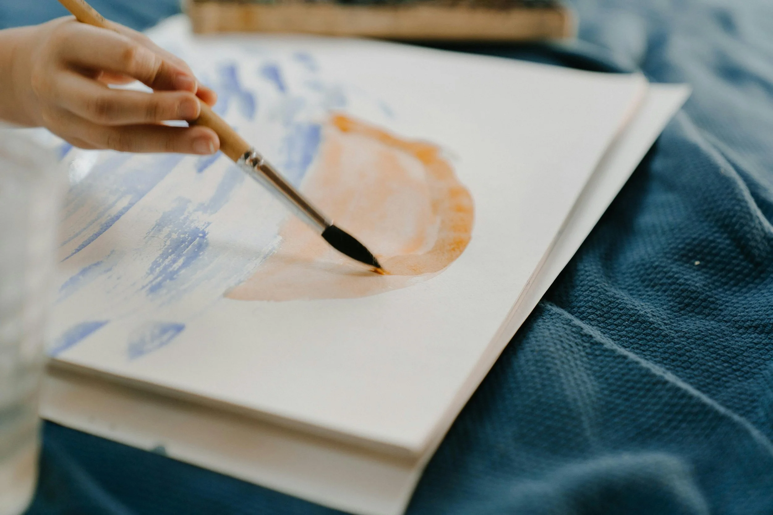 Person painting with watercolor on paper, using a paintbrush, creating abstract shapes in blue and orange.