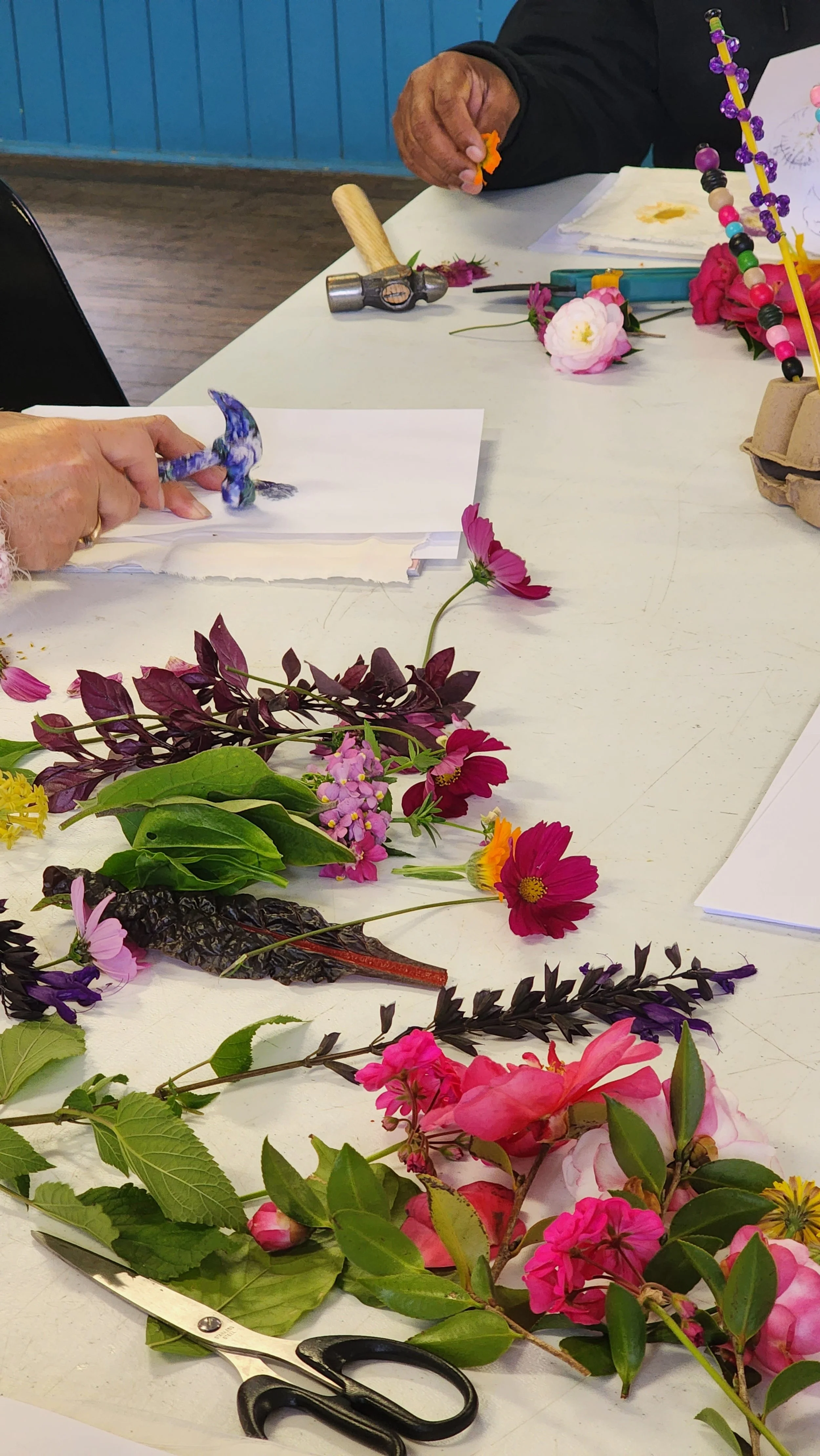 People working on floral arrangements with various flowers, leaves, scissors, and other craft tools on a white table.
