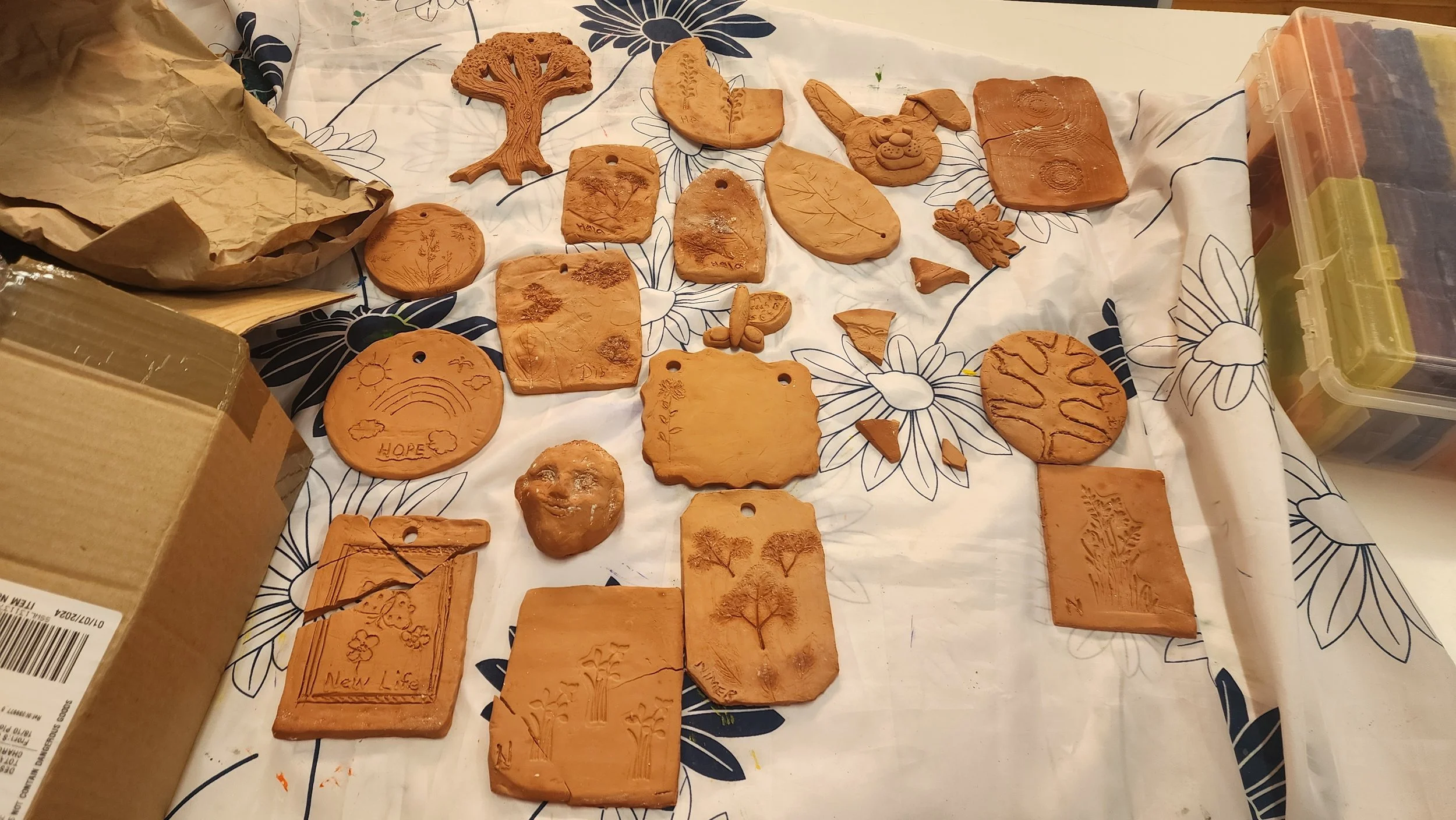 Clay plaques with carved images of trees, animals, and nature scenes on a table covered with floral patterned paper.