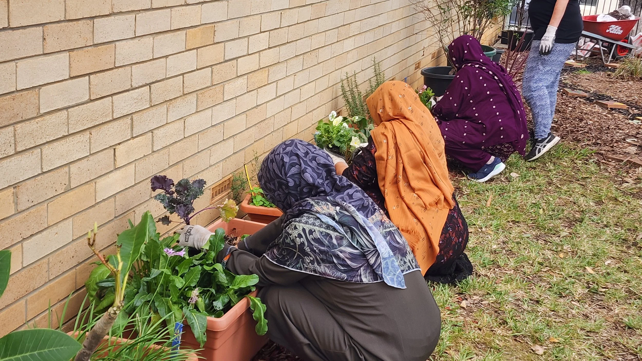 Three women wearing colorful headscarves and casual clothing planting flowers in rectangular pots along a brick wall in a garden.