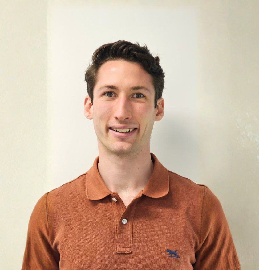 A photo of Rees Hopper - a young man with short brown hair and blue eyes smiling, wearing a brown polo shirt with a small navy emblem of a dog on the left chest.