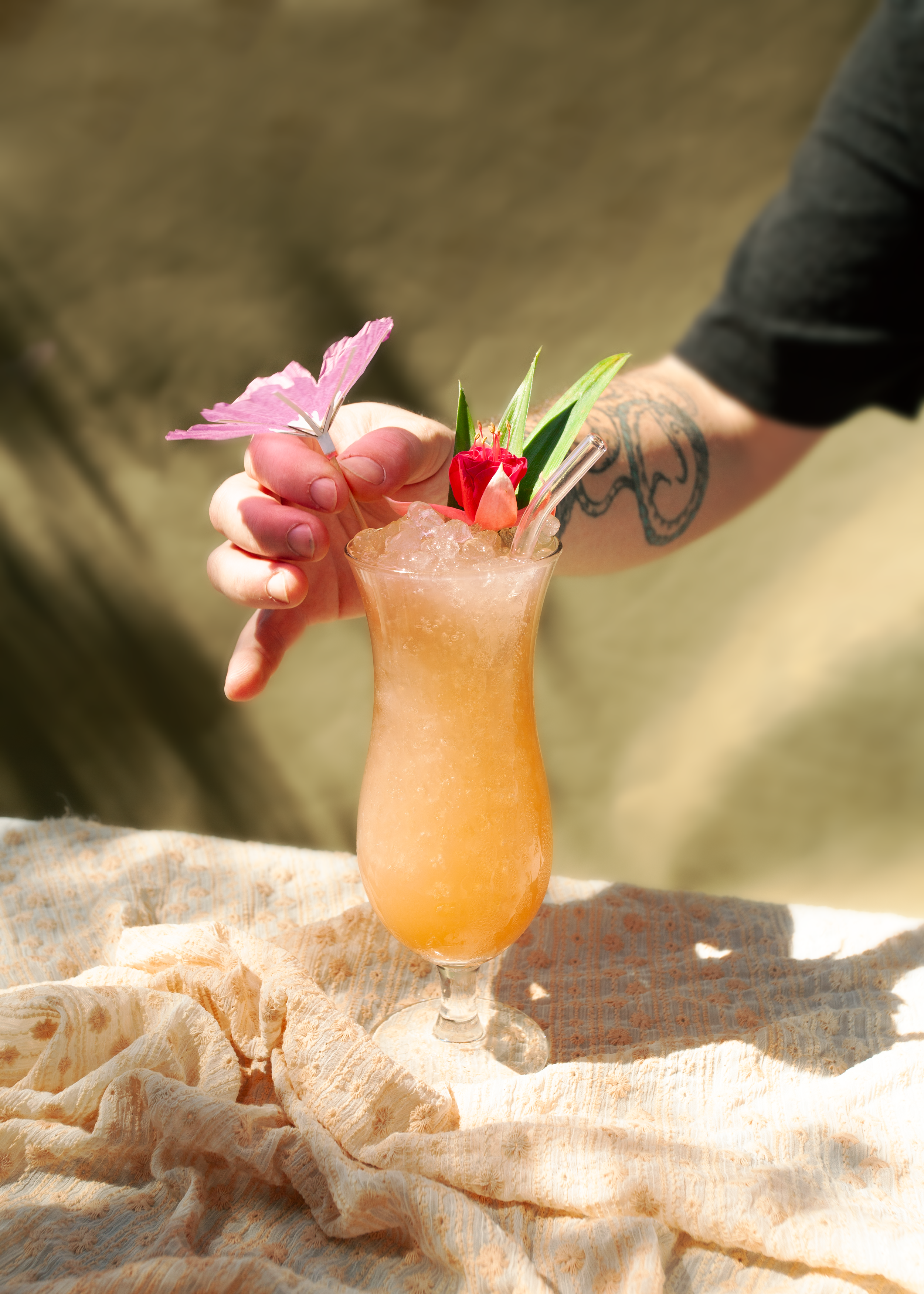 Frosty orange cocktail garnished with flowers and herbs, held by a tattooed hand.