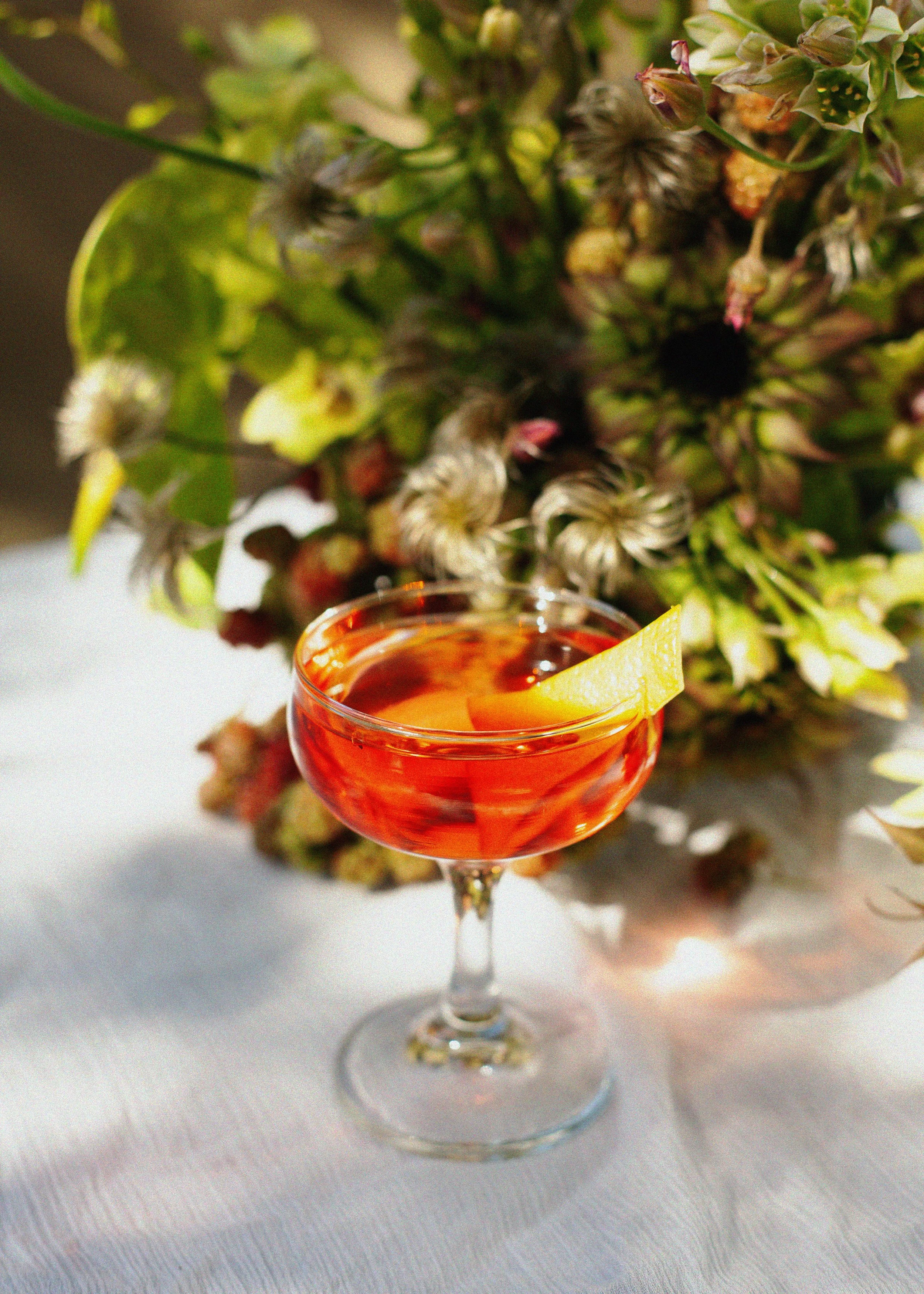 Close-up of an orange cocktail with a lemon wedge, surrounded by green foliage and dried flowers.