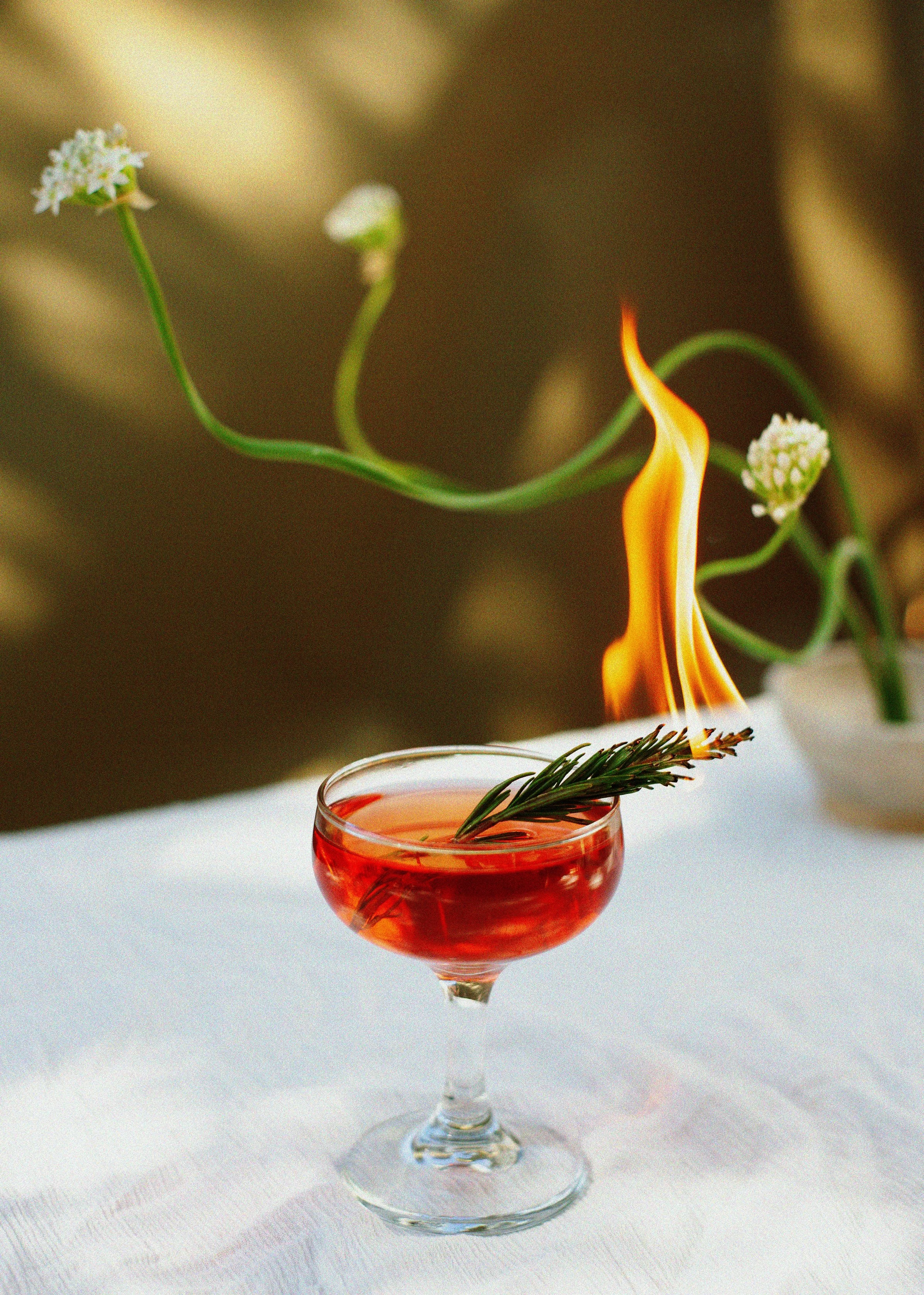 Red cocktail in a coupe glass garnished with rosemary, surrounded by white flowers and warm candlelight.