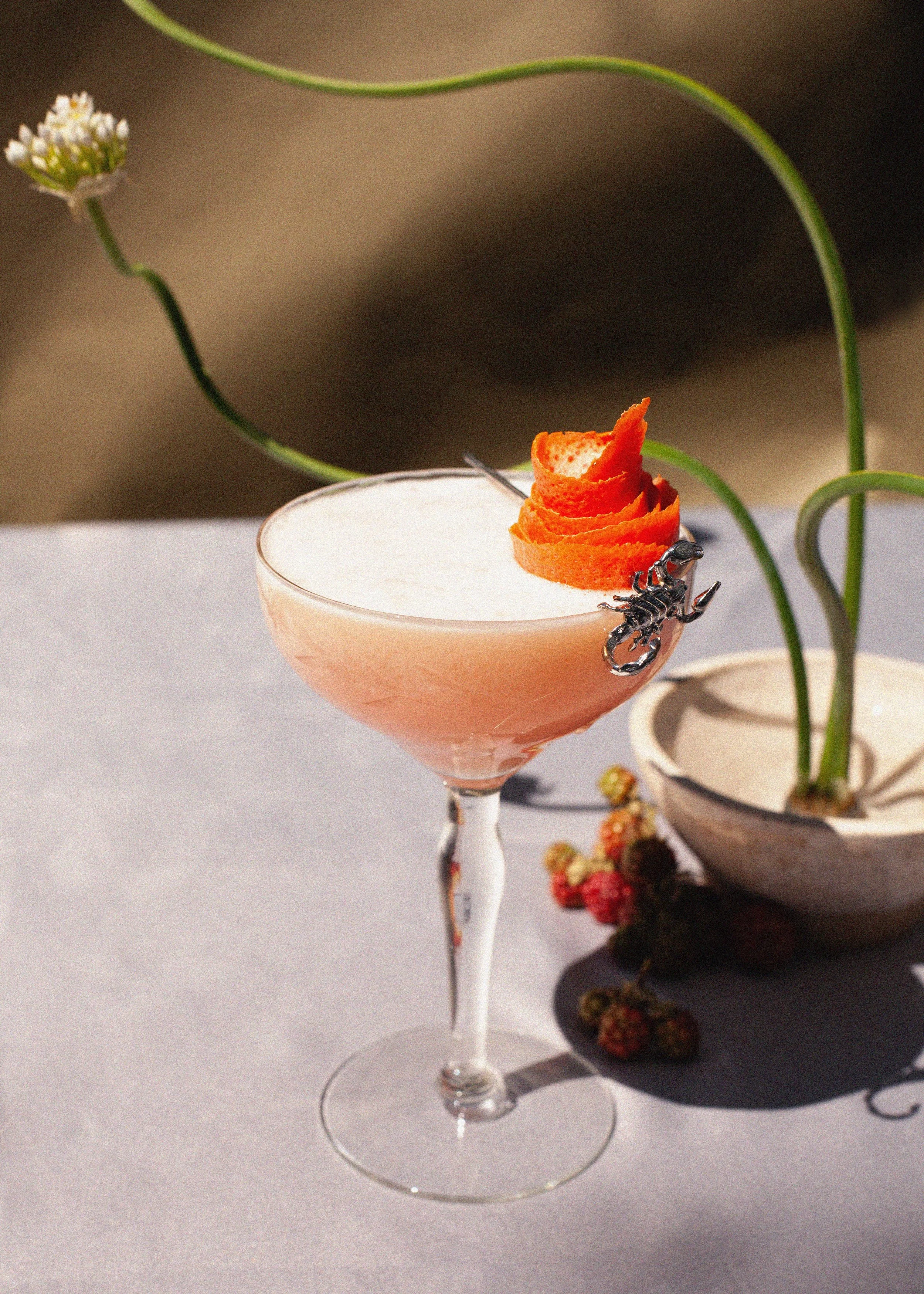 Pink cocktail in a coupe glass garnished with a curled orange peel and black scorpion decoration, beside a black plate of hors d'oeuvres