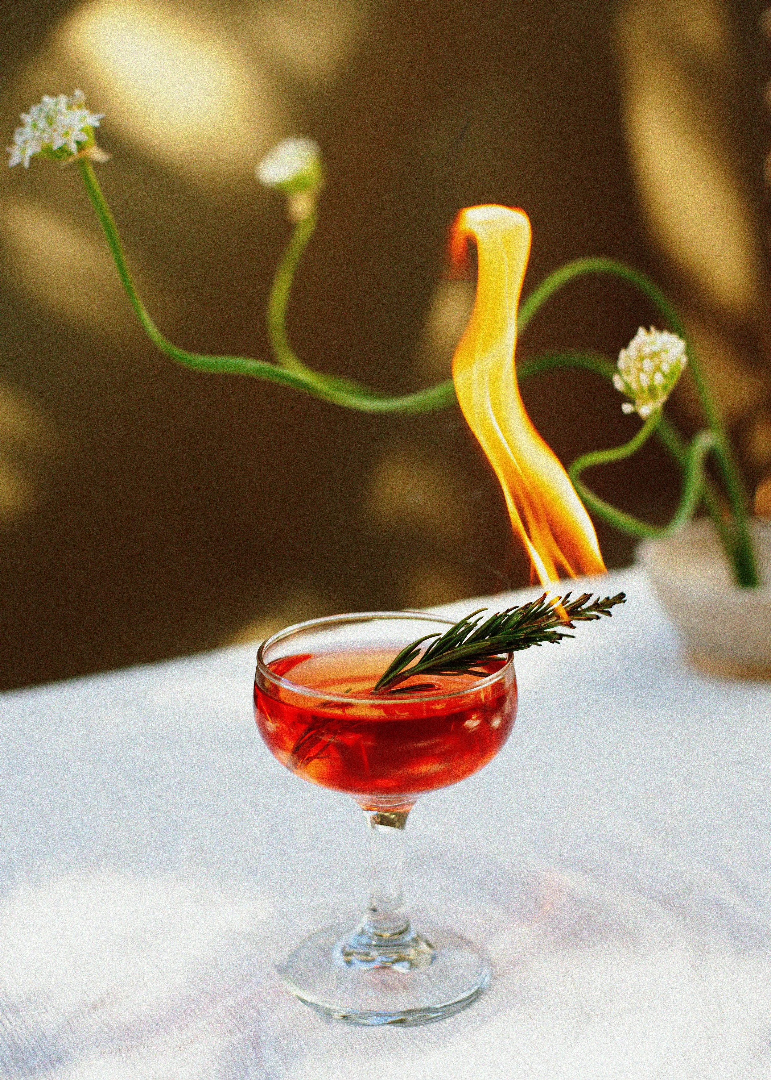 Red cocktail in a stemmed glass garnished with rosemary, surrounded by white flowers and warm candlelight.