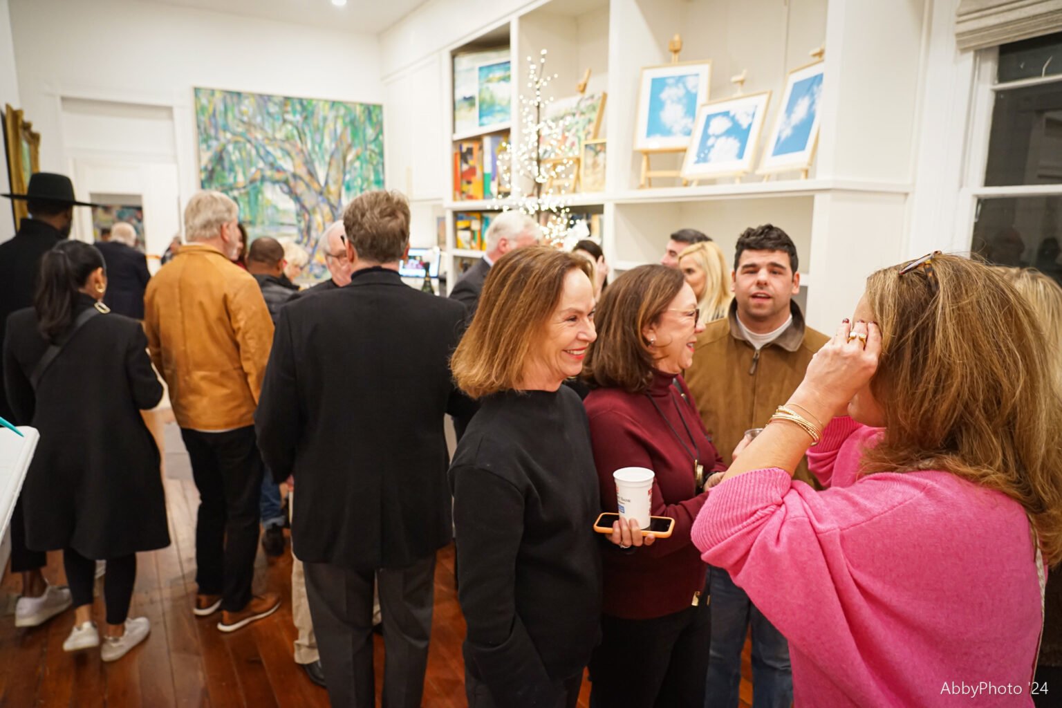 Group of people socializing at an art gallery or event, with paintings hanging on the walls and a Christmas tree in the background.