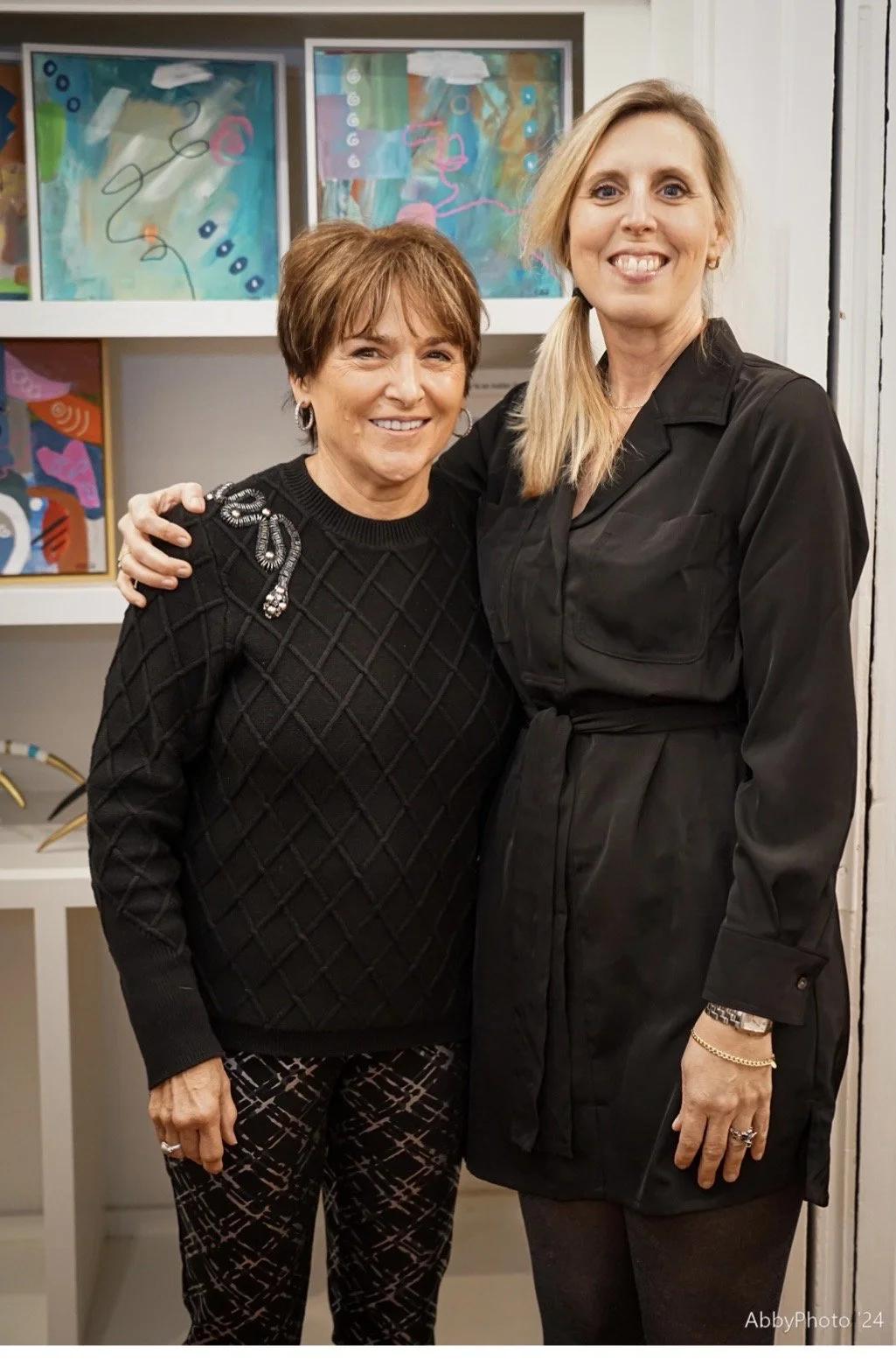 Two women smiling and standing side by side in front of abstract colorful artwork. One woman has short brown hair and is wearing a black sweater with a decorative pin, patterned pants, and earrings. The other woman has long blond hair and is wearing 