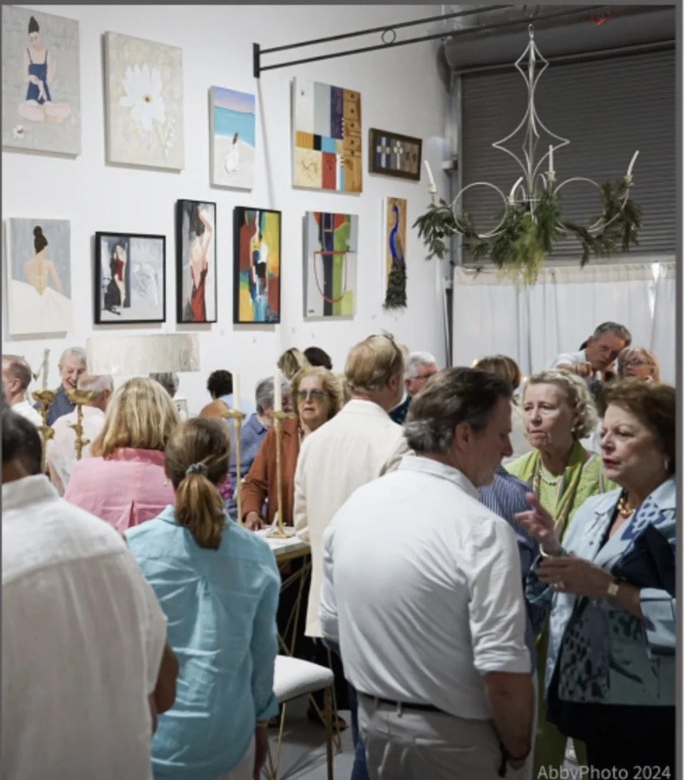A gathering of mostly older adults socializing in an art gallery or art exhibition space with white walls decorated with colorful paintings and artworks, a chandelier with greenery hanging from it, and a large window with curtains in the background.