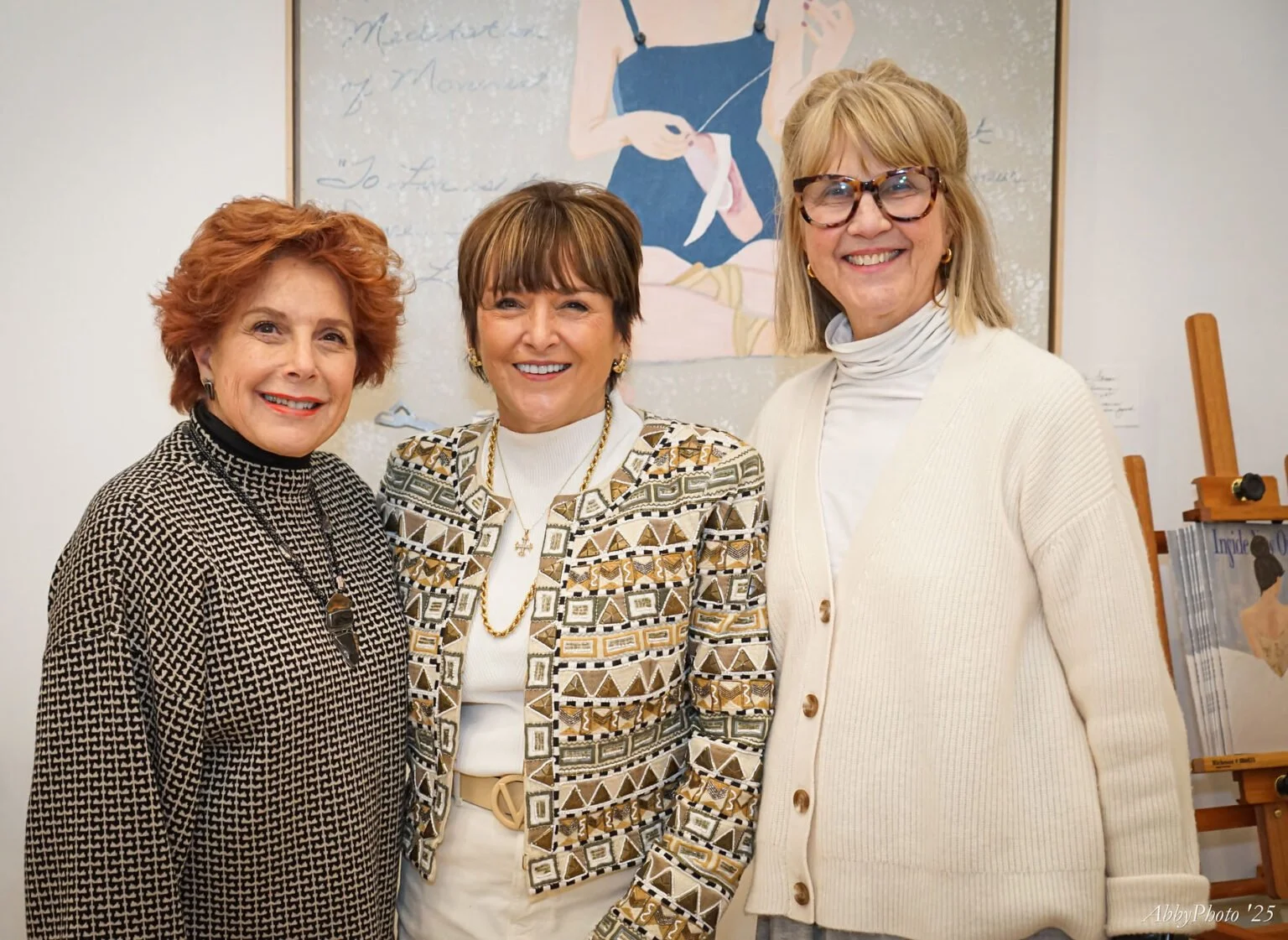 Three women standing together indoors, smiling at the camera. The woman on the left has red hair and is wearing a black and white patterned blazer. The woman in the middle has short brown hair, wearing a patterned blazer with gold accents, a white tu