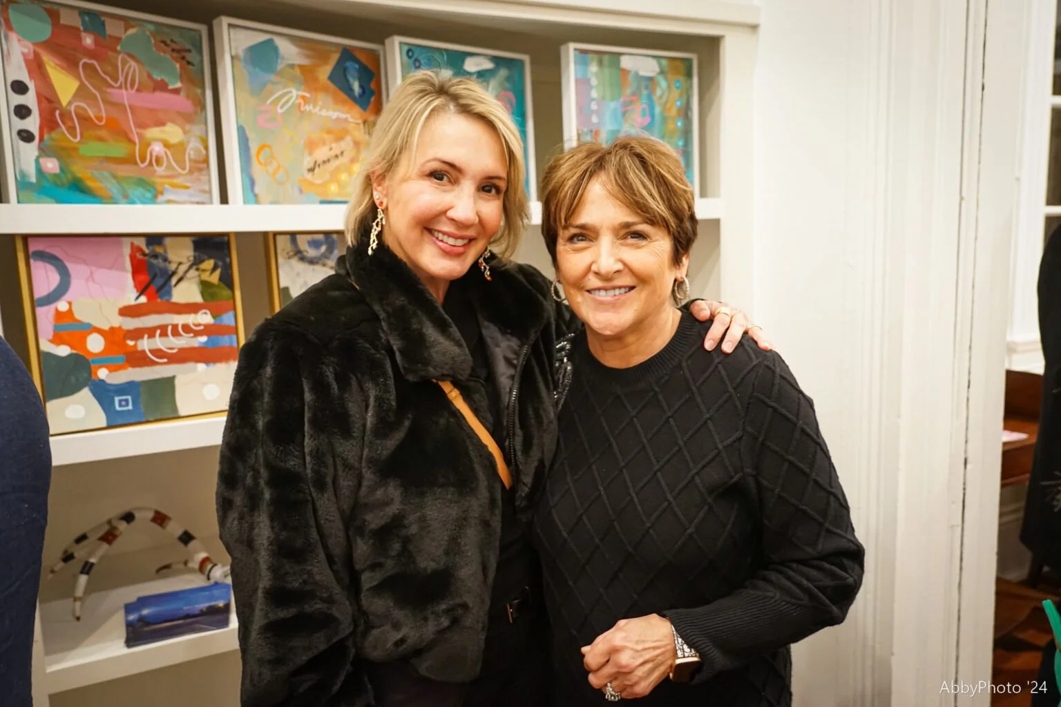 Two smiling women pose together indoors, one with blonde hair and the other with short brown hair, with colorful abstract art on the wall behind them.
