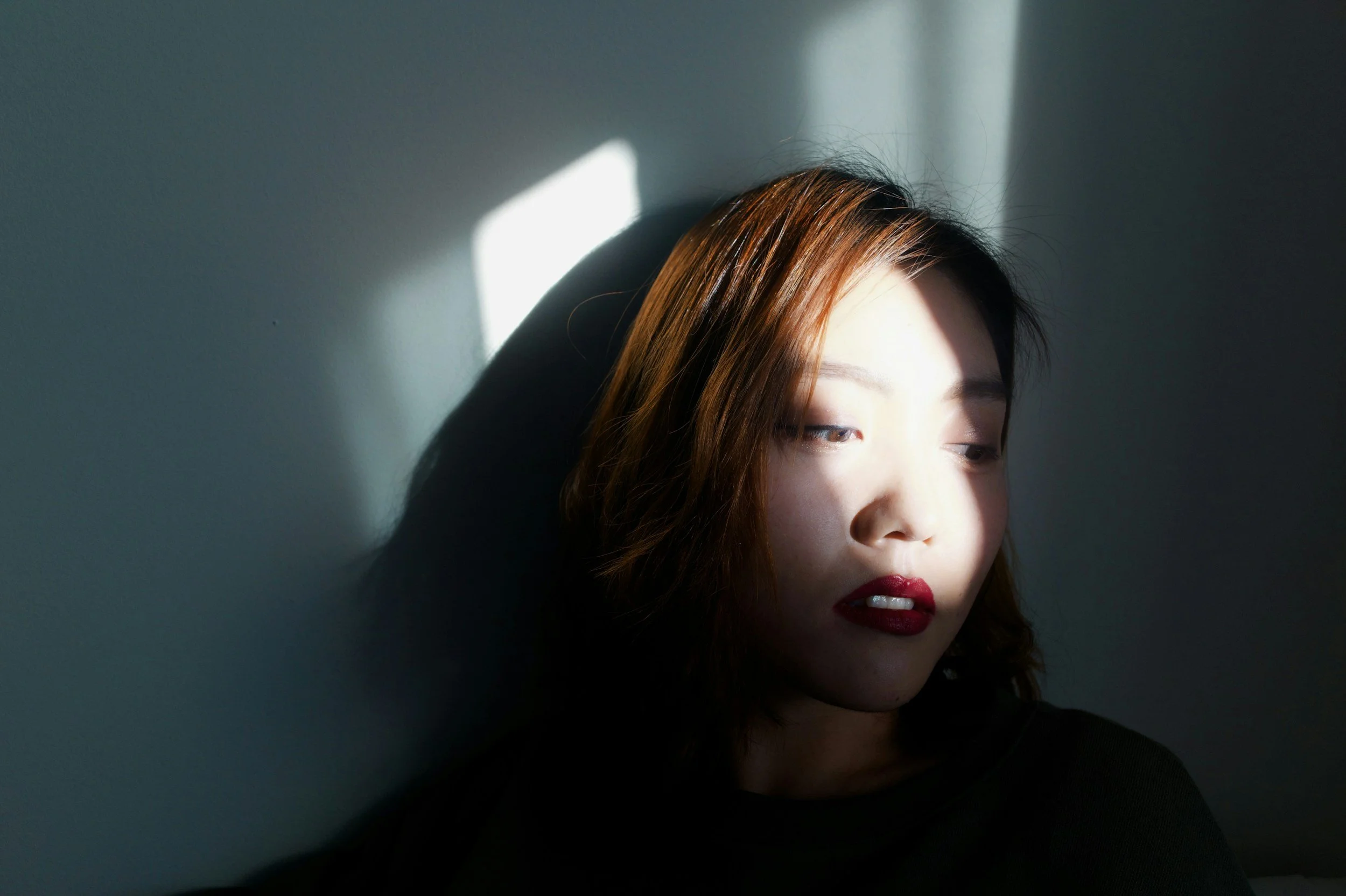A young woman with medium-length brown hair and red lipstick is facing a wall with sunlight casting shadows on her face.