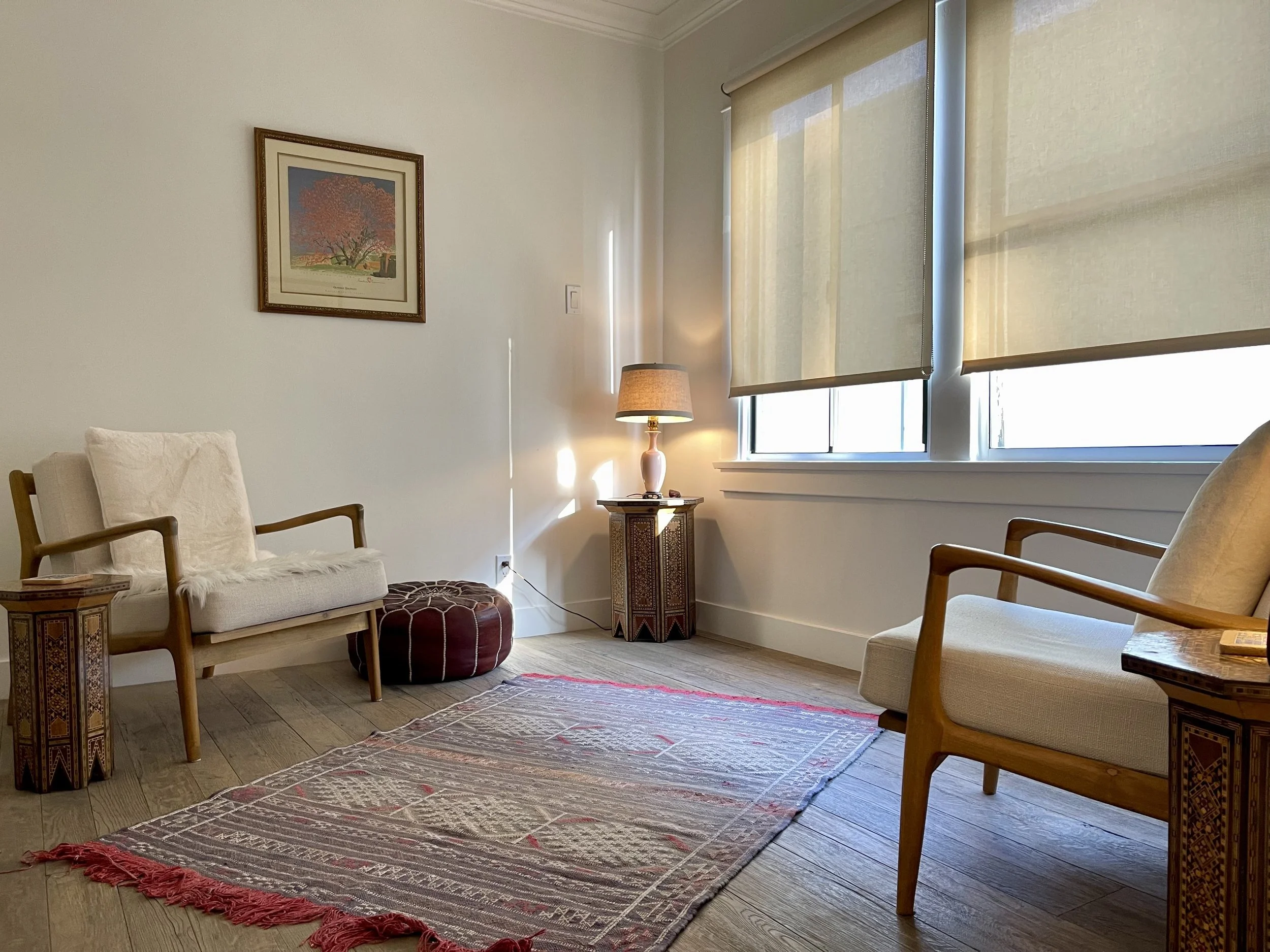 A cozy living room with two beige armchairs with wooden arms, a wooden side table with intricate design, and a round red Ottoman in the corner. There is a colorful patterned rug on the light wooden floor, a framed picture of a tree with orange leaves hanging on the white wall, and a window with beige blinds allowing sunlight to softly illuminate the space. A lamp with a beige shade sits on a decorative wooden stand next to the window.