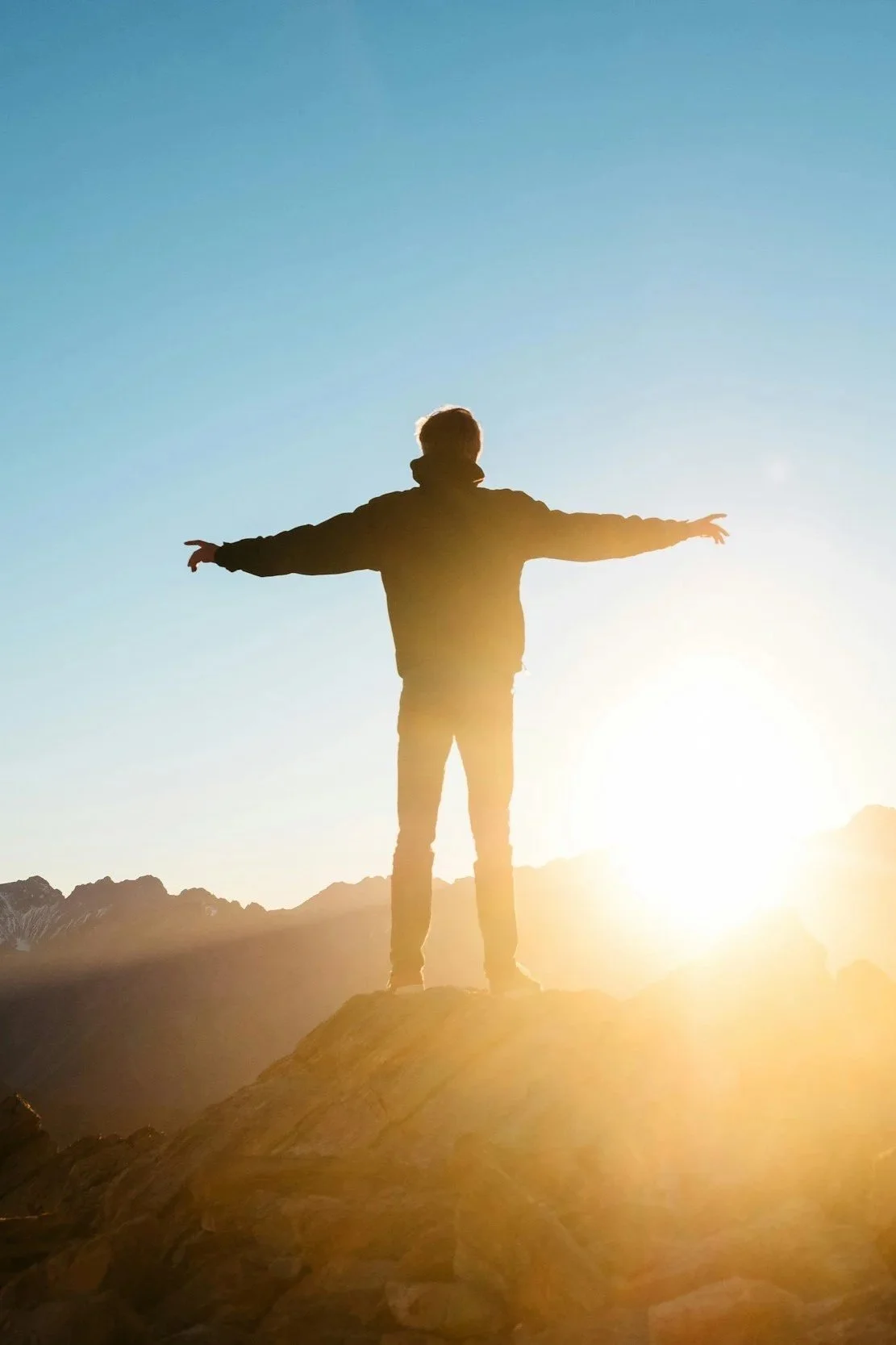 Person standing on a rock with arms outstretched during sunset or sunrise, with mountains in the background and clear sky.
