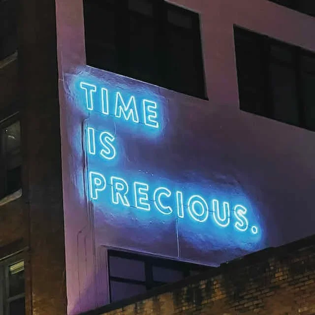 Neon sign on building wall that reads "Time is precious" in blue illuminated letters