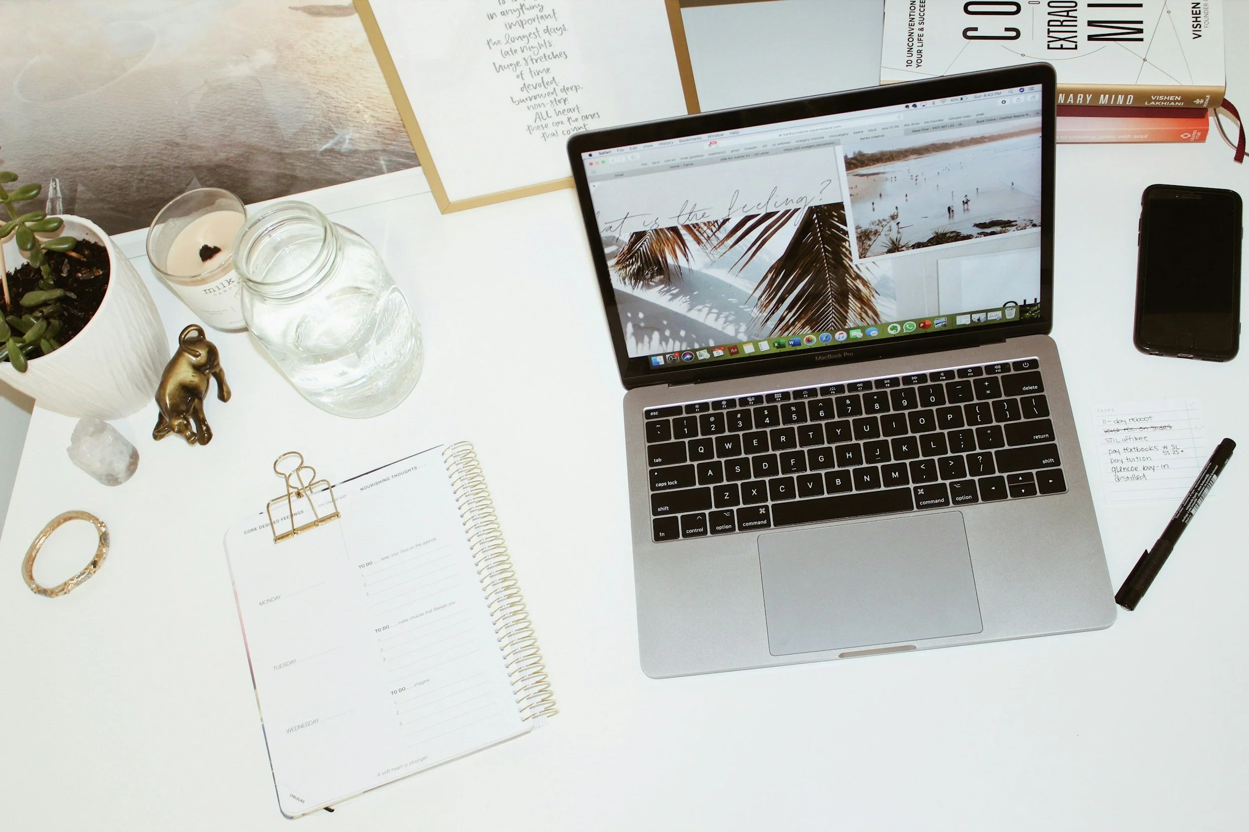 A workspace with a laptop displaying a beach scene, a smartphone, a notepad with handwritten notes, a pen, a planner, a glass of water, a candle, a small plant, a decorative elephant figurine, a bracelet, and a jar of water on a white desk.