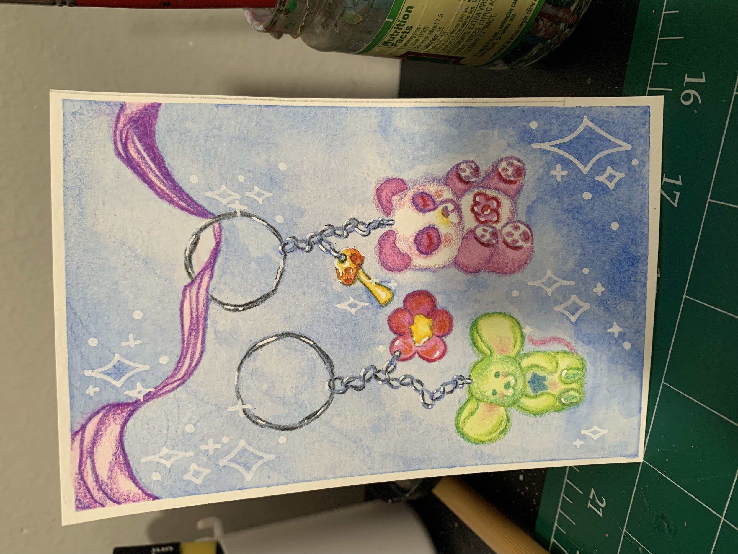 Illustration of two colorful cute bunny-shaped keychains, one pink and one green, connected by chains, set against a pastel blue background with white sparkles and a purple swirl.