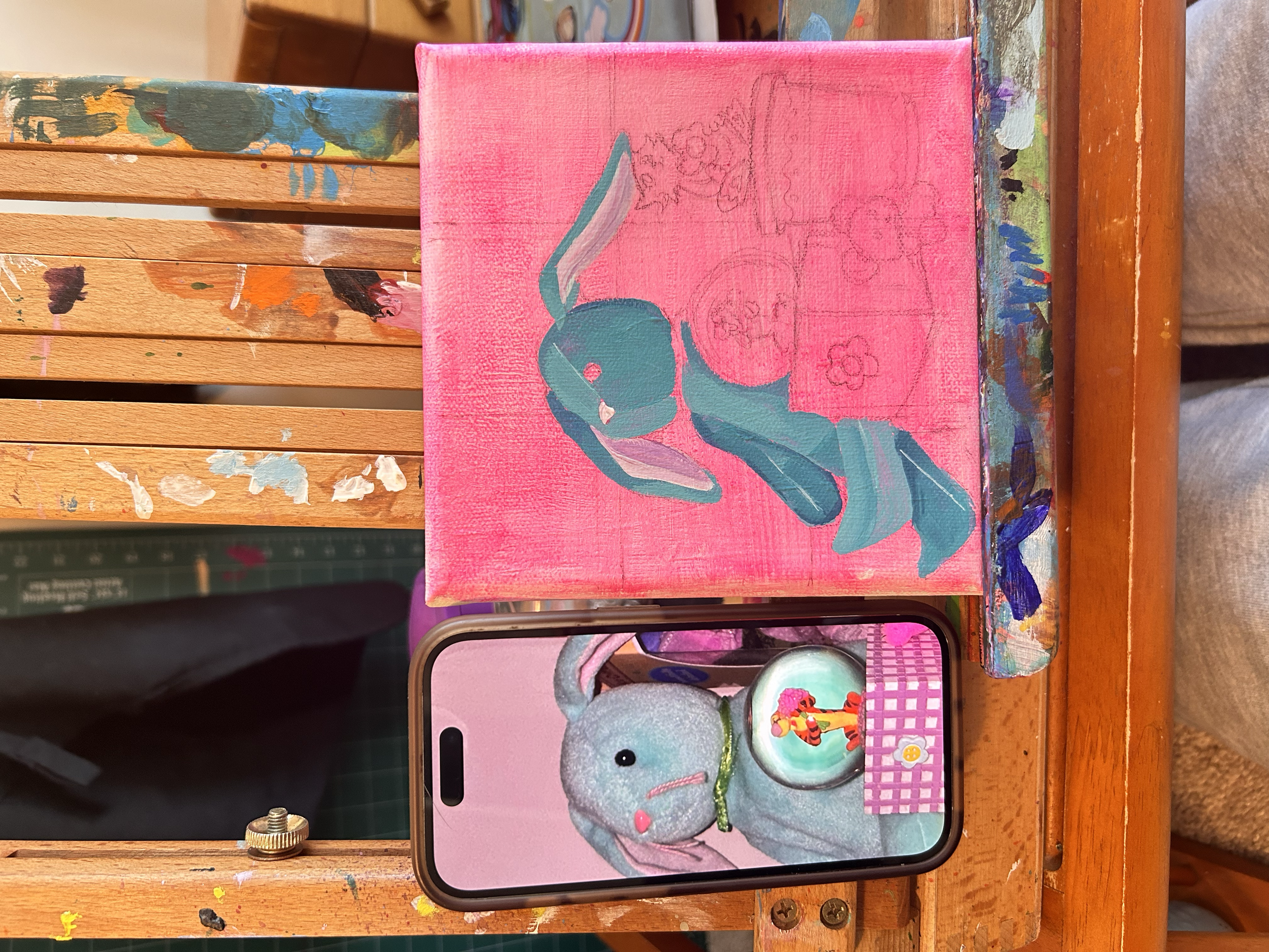 A pink canvas with an unfinished painting of a blue bunny on a pink background. In front of the canvas, there is a smartphone displaying a digital photo of a stuffed gray bunny with a silver ball marked with a picture of a colorful toy figure, placed