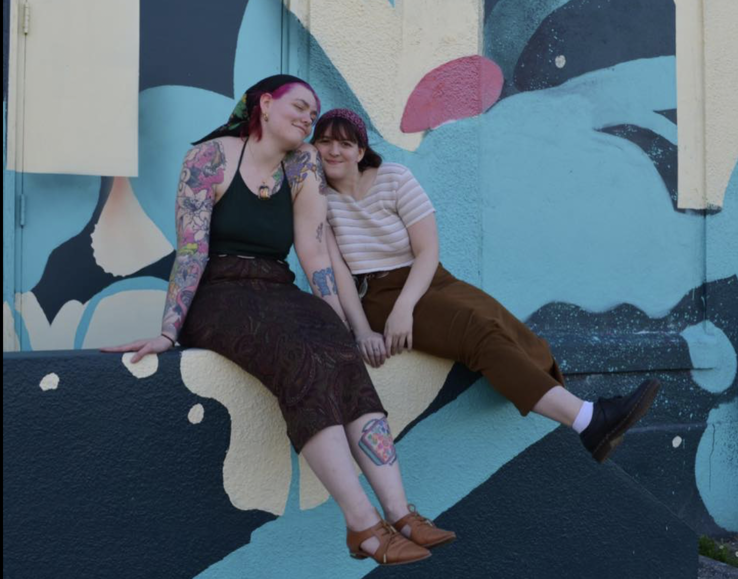 Artist couple Kayla Bugs and Ginni ghost sitting on a ledge with a blue and beige mural behind them.