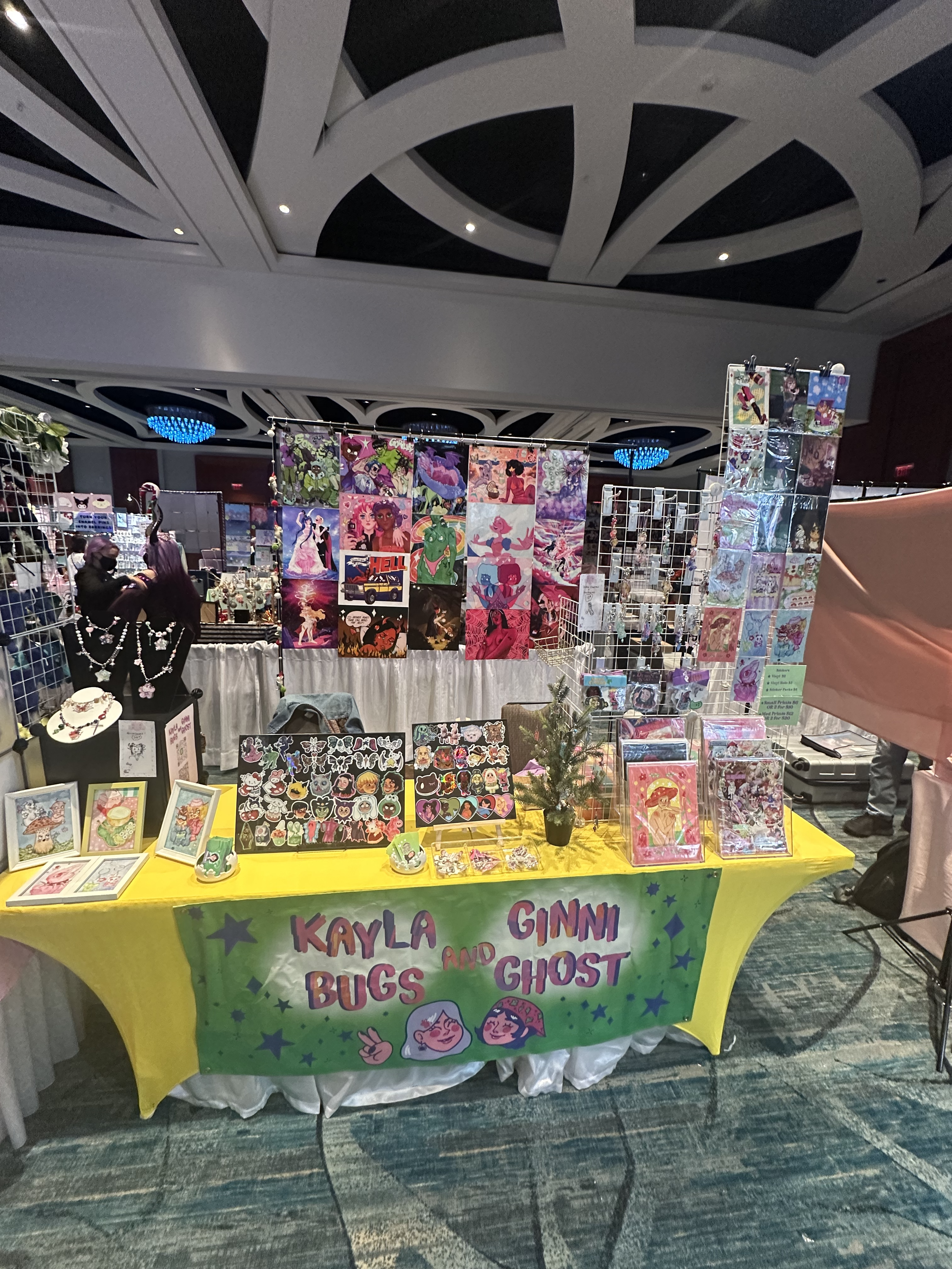 Vendor booth at a convention, selling colorful fan art, patches, jewelry, and accessories. The booth has a yellow tablecloth with a banner that says "Kayla Bugs and Ginni Ghost," featuring cartoon characters. Background includes other booths and deco