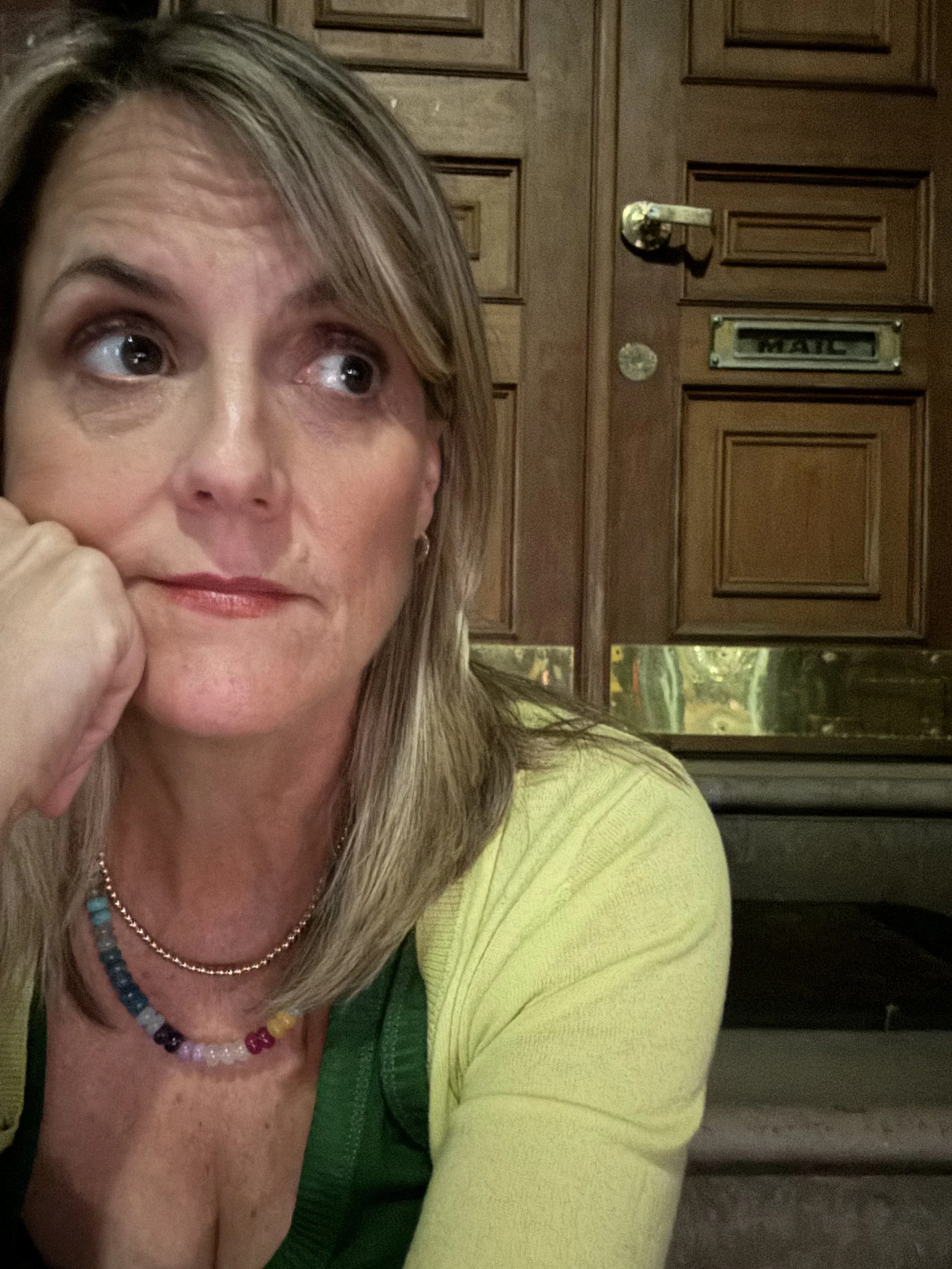 A woman with blonde hair resting her face on her hand, looking to the side, sitting indoors with a wooden door in the background.