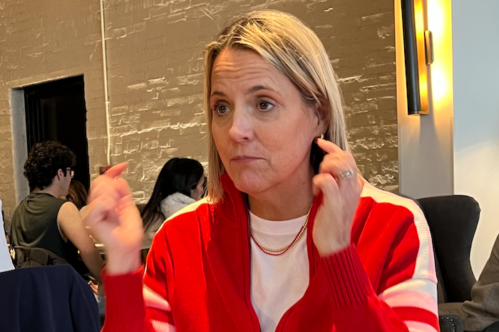 A woman with blonde hair wearing a red and white jacket, sitting in a restaurant or cafe, touching her ear with her right hand and gesturing with her left hand, with other people in the background.
