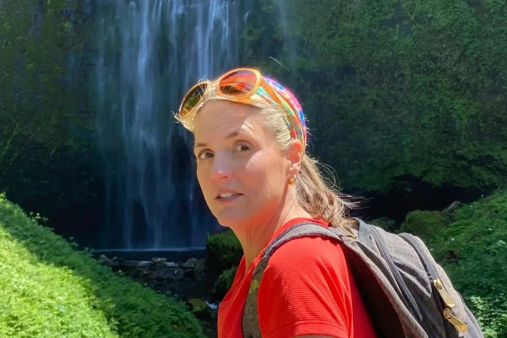 A woman with blonde hair, wearing sunglasses and a red shirt, hiking with a backpack in a lush green area, with a waterfall in the background.