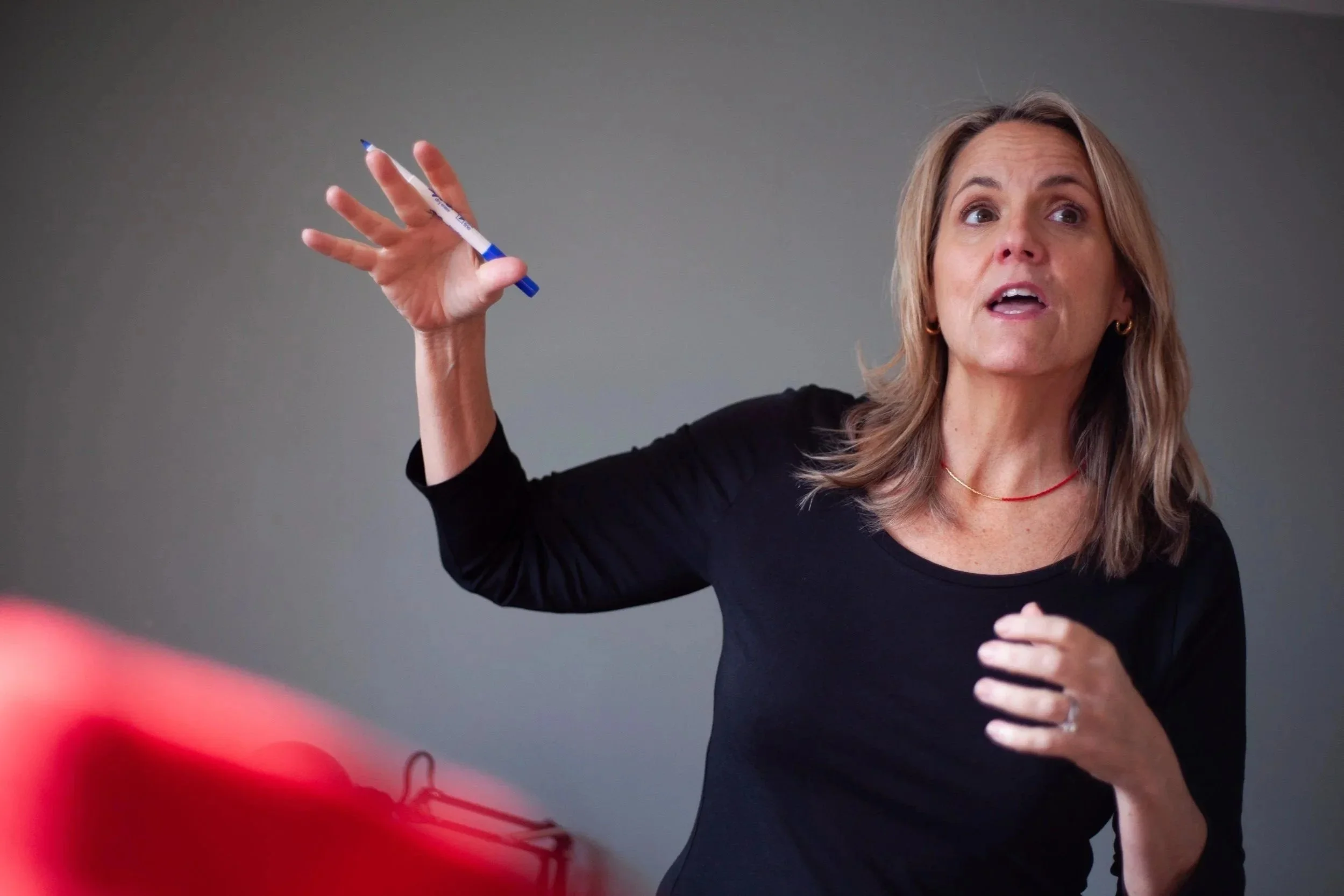 A woman with shoulder-length blonde hair speaking and gesturing with her right hand, holding a blue and white marker, and wearing a black shirt and a red necklace.