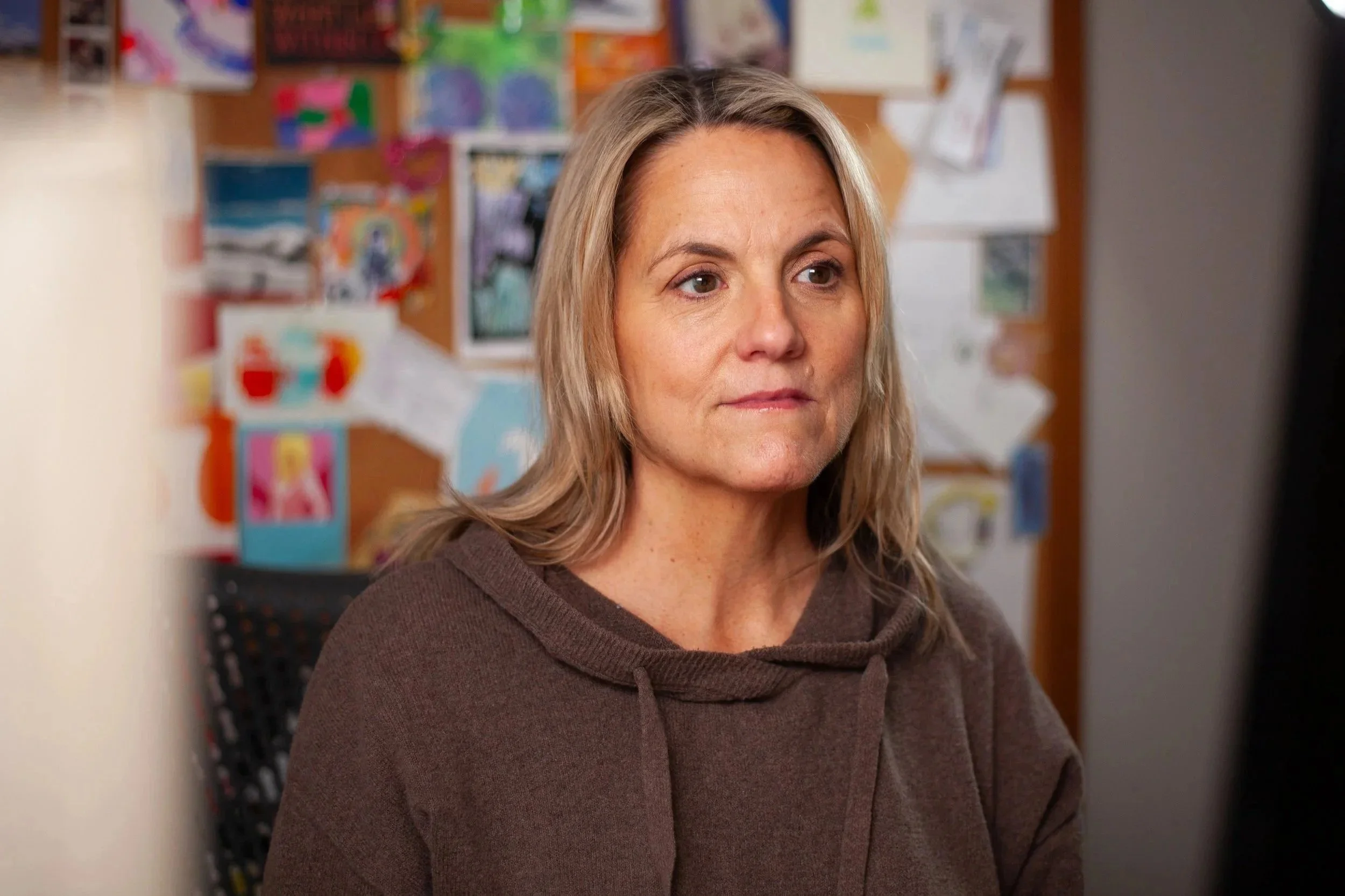 A middle-aged woman with blonde hair looking at a screen, wearing a brown hoodie, with a colorful bulletin board in the background.