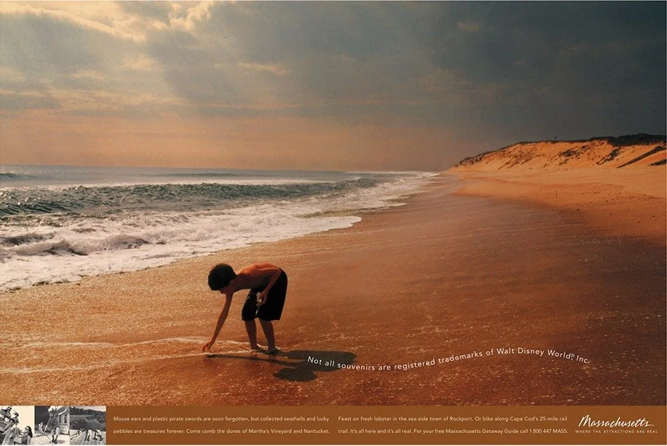 A young boy is playing on a sandy beach near the shoreline, with waves approaching and a distant cliff in the background during sunset.