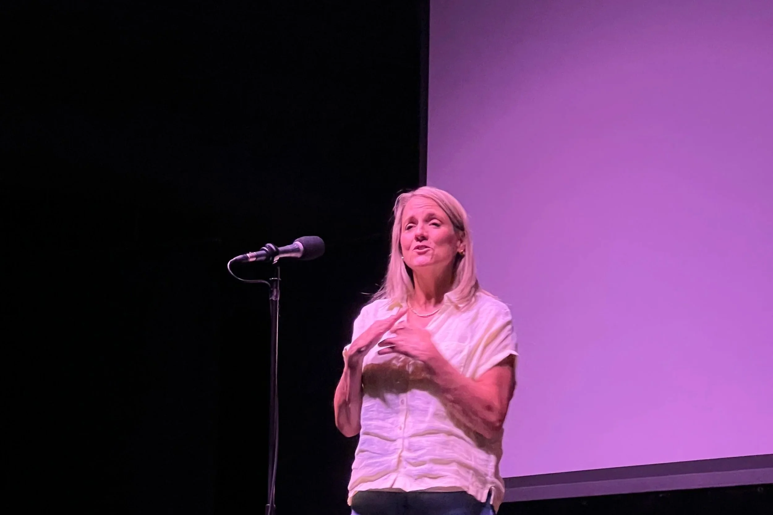 A woman with blonde hair speaking into a microphone on a stage with a purple and black background.