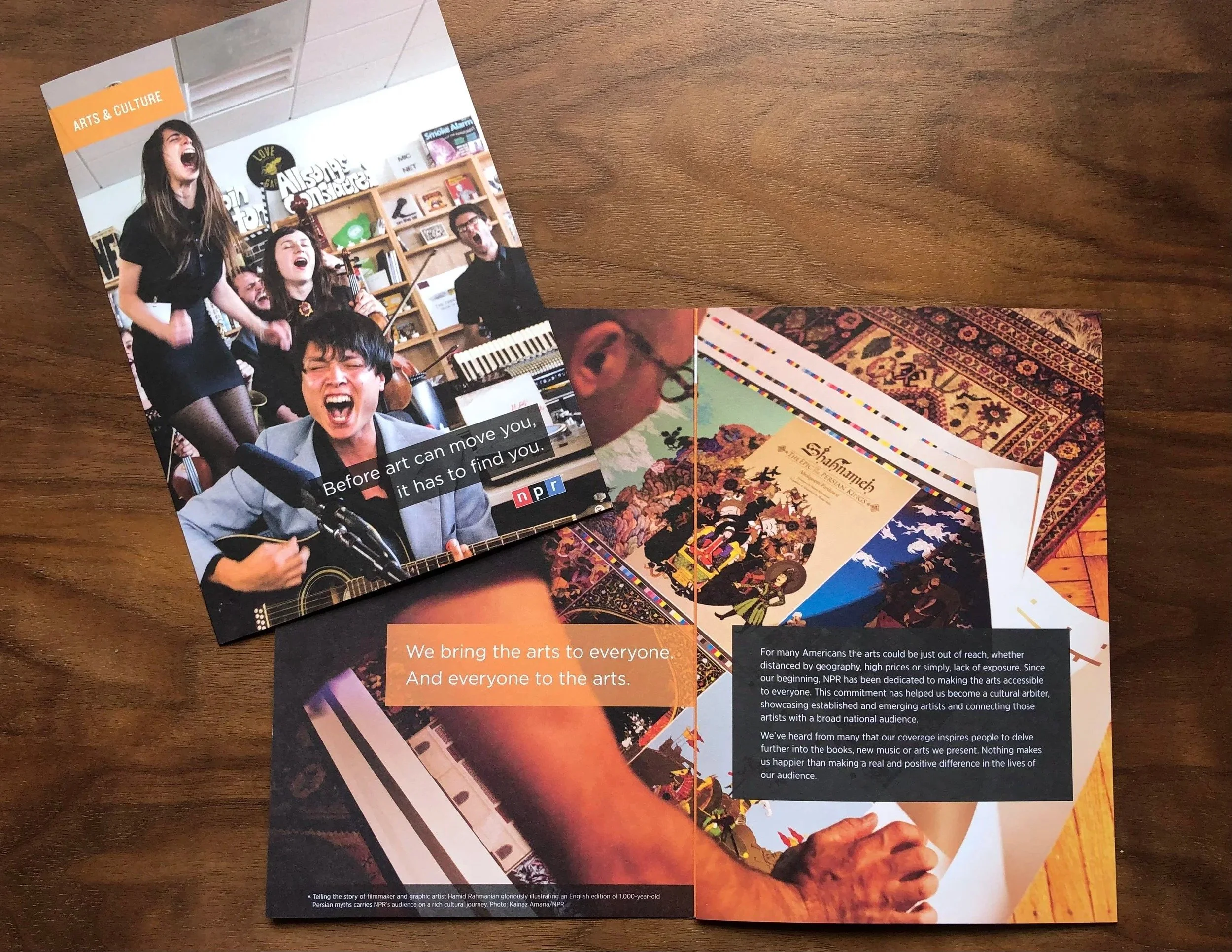 A brochure with two pages open on a wooden surface. The left page features a vibrant photo of a group of people singing and playing musical instruments, with subtitles and the NPR logo. The right page contains images of artwork, a person reading a book, and text about making arts accessible to everyone.