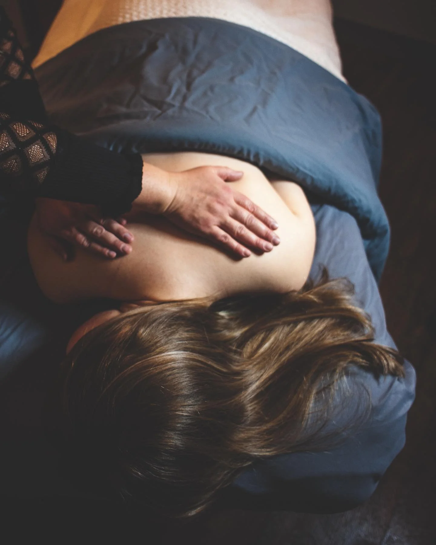 A person receiving a massage on their back while lying on a bed with a gray blanket, with the person's head turned to the side and brown hair visible.