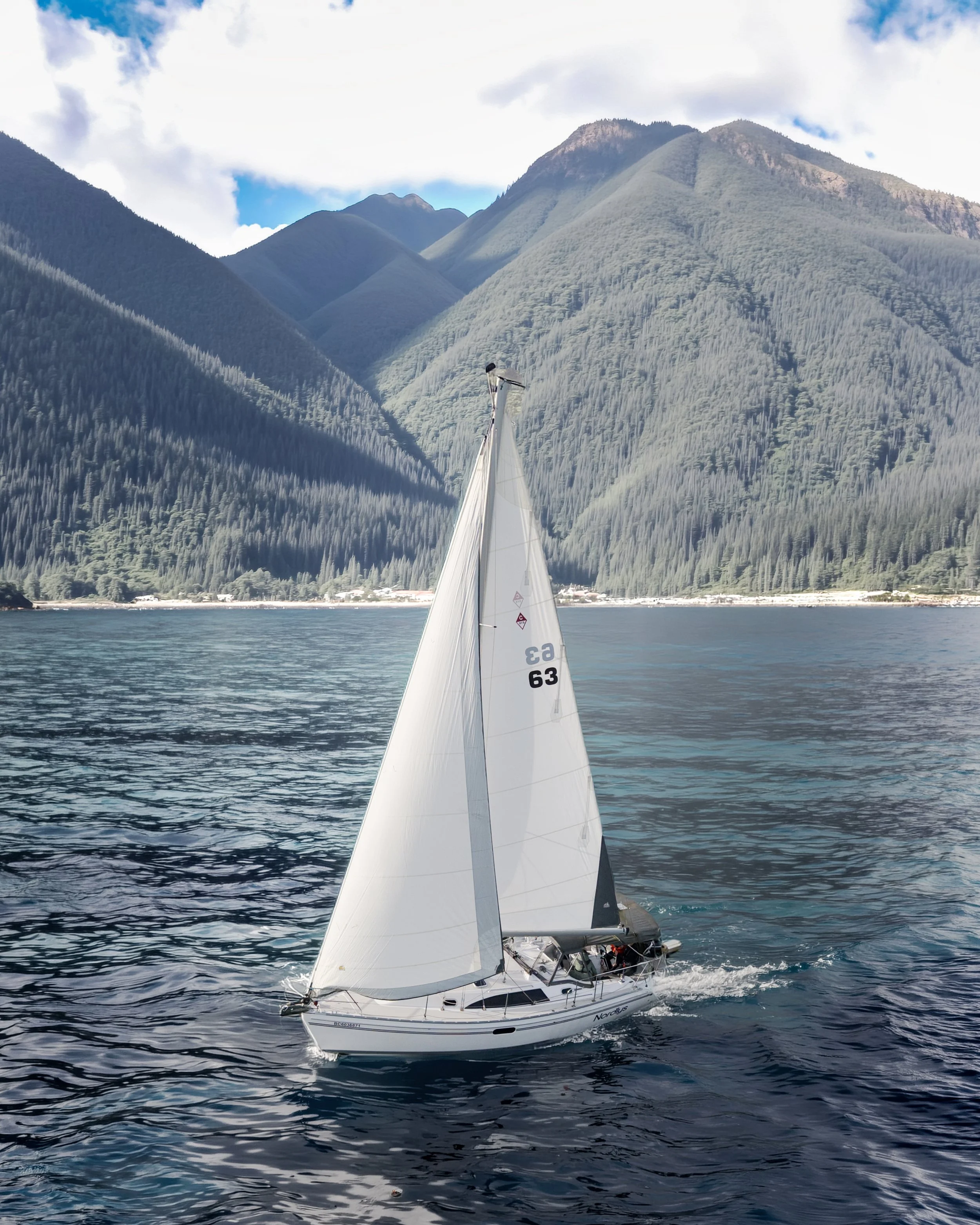 A sailboat with white sails sailing on a body of water surrounded by green mountains under a partly cloudy sky.