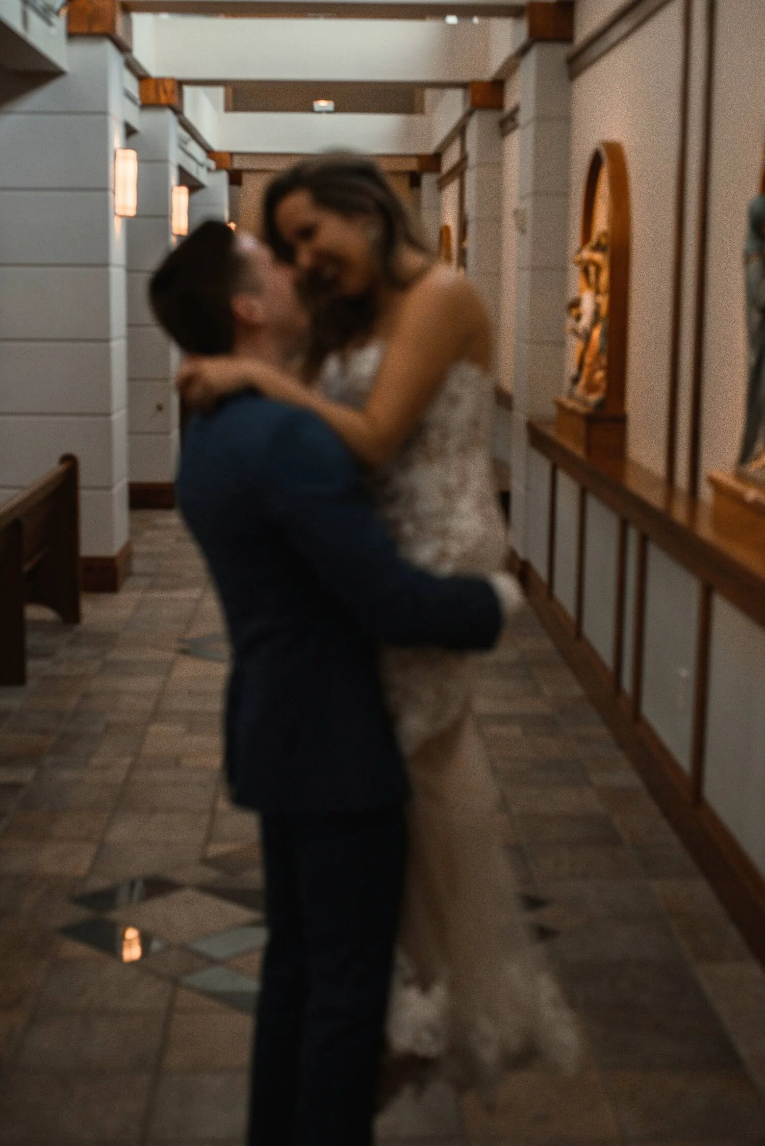A man and woman celebrating at a wedding, with the man lifting the woman in his arms and kissing her, in a warmly lit indoor venue.