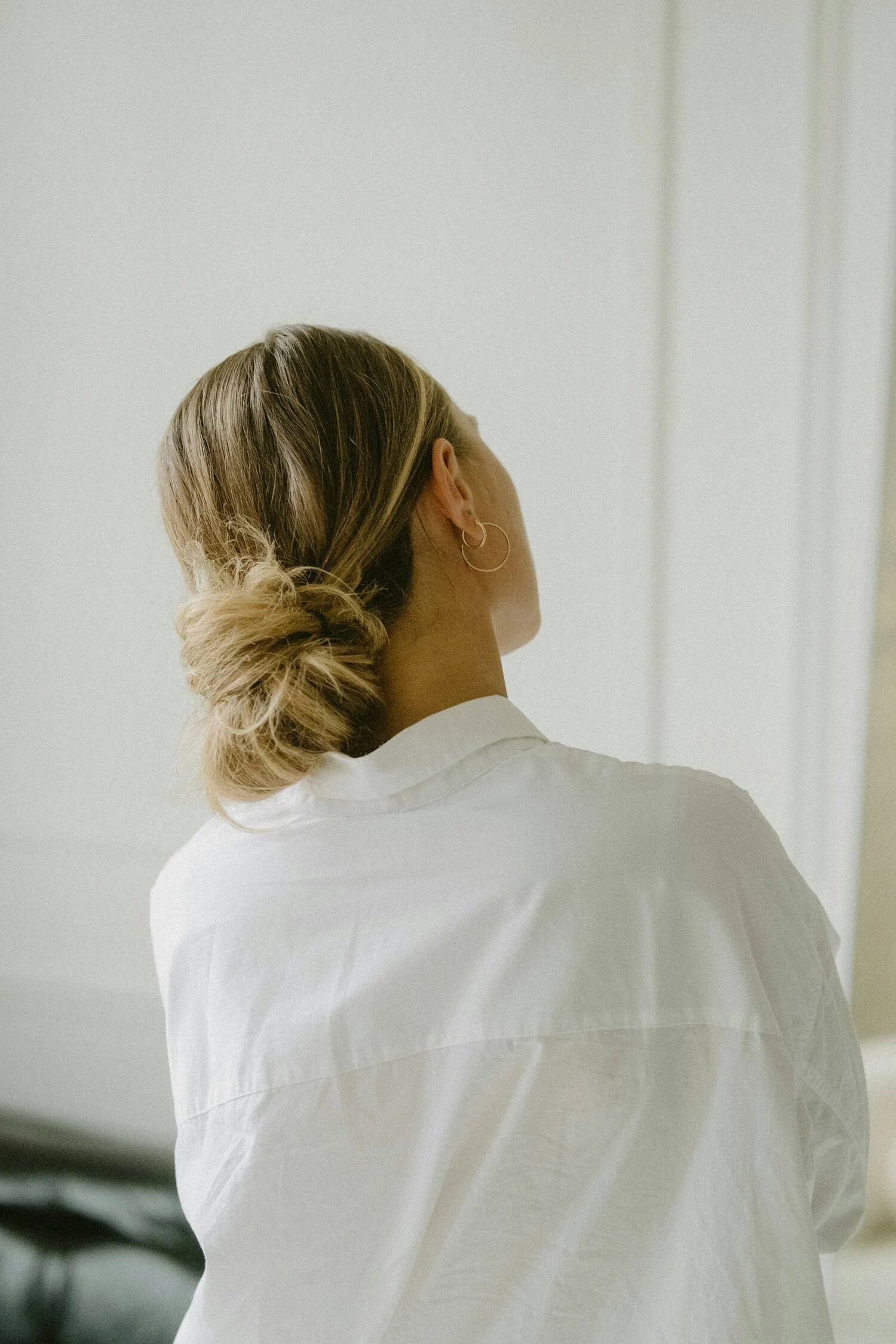 A woman with blonde hair tied in a messy bun, wearing hoop earrings and a white shirt, facing away from the camera in a minimal interior setting.