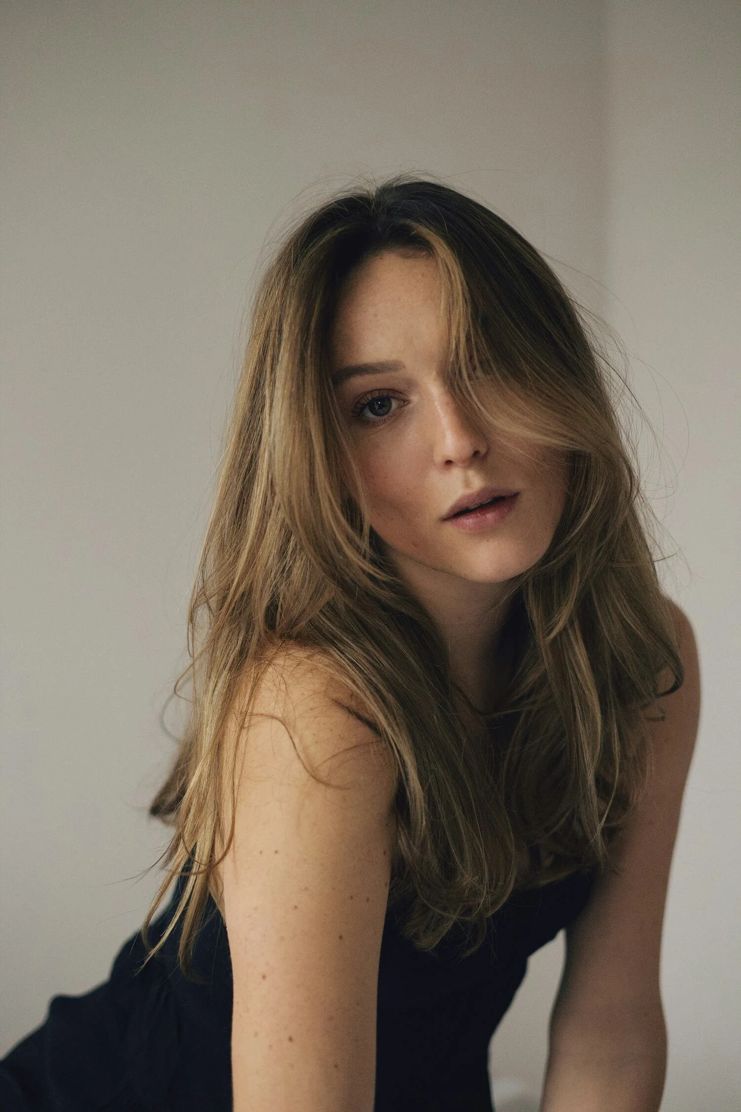 A woman with long, wavy brown hair, partially covering her face, wearing a black sleeveless top, looking at the camera with a neutral expression.