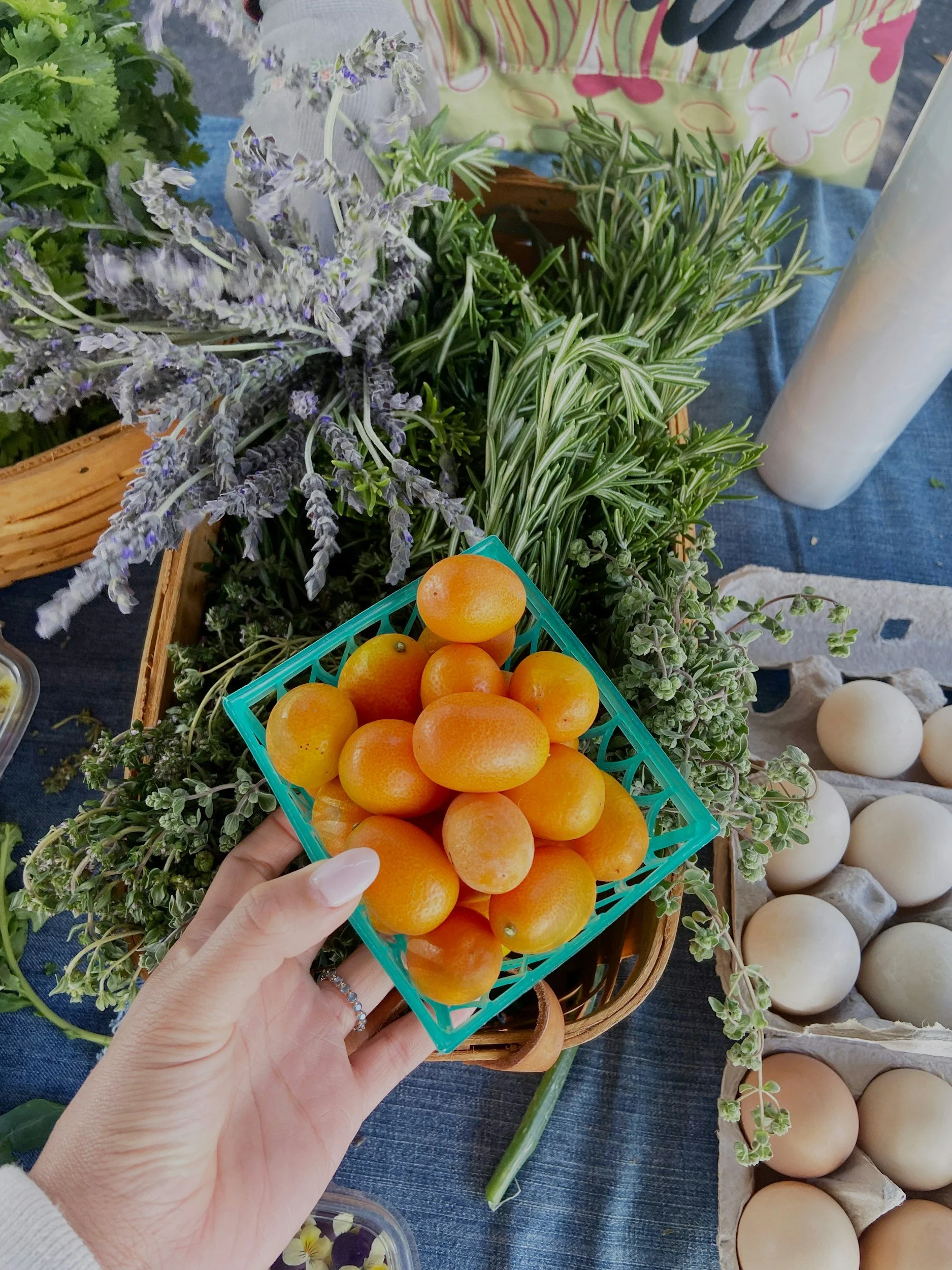 Person holding a small basket of yellow tomatoes at a farmer's market with fresh herbs and eggs on display.