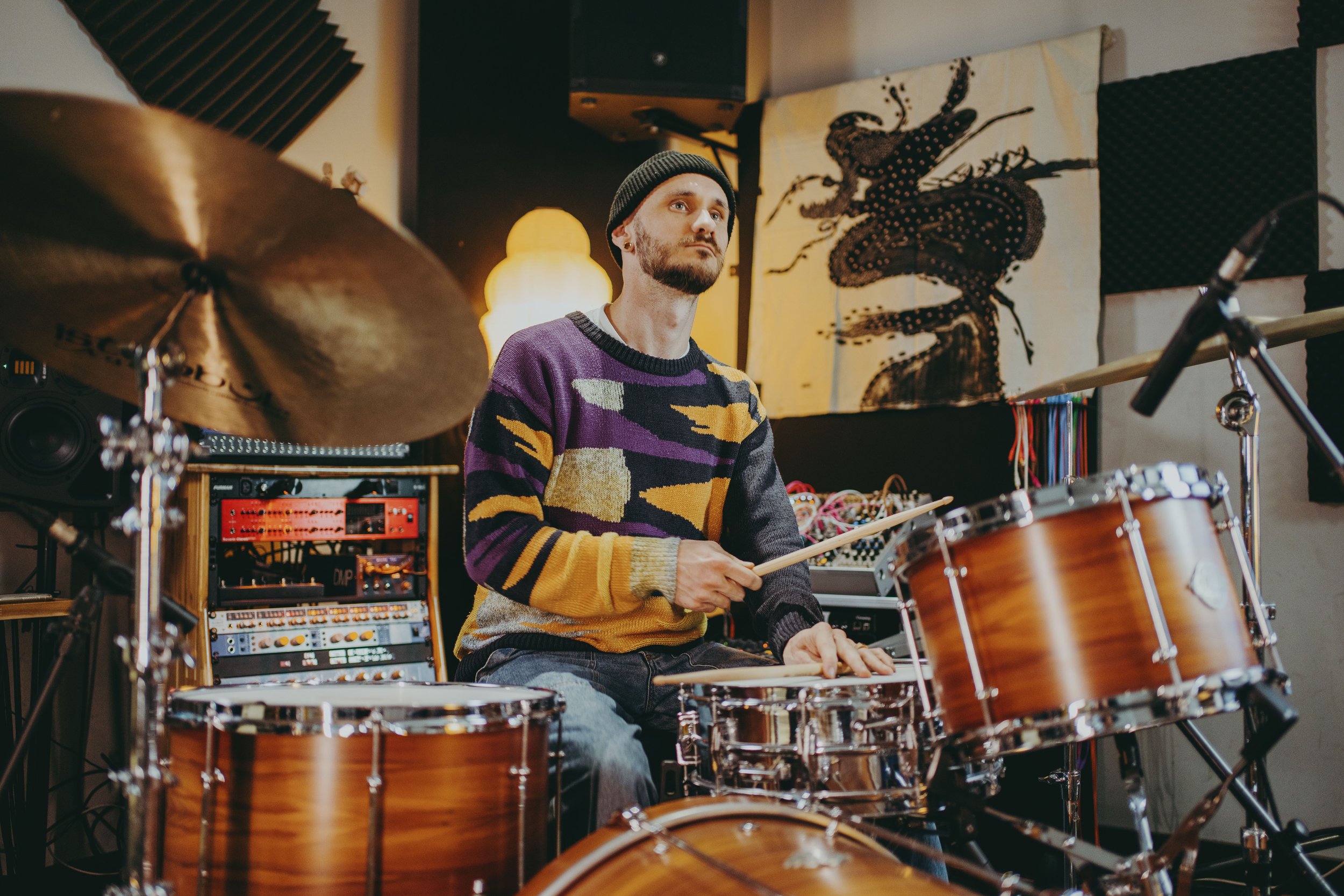 A man wearing a colorful sweater and beanie playing drums in a music studio.