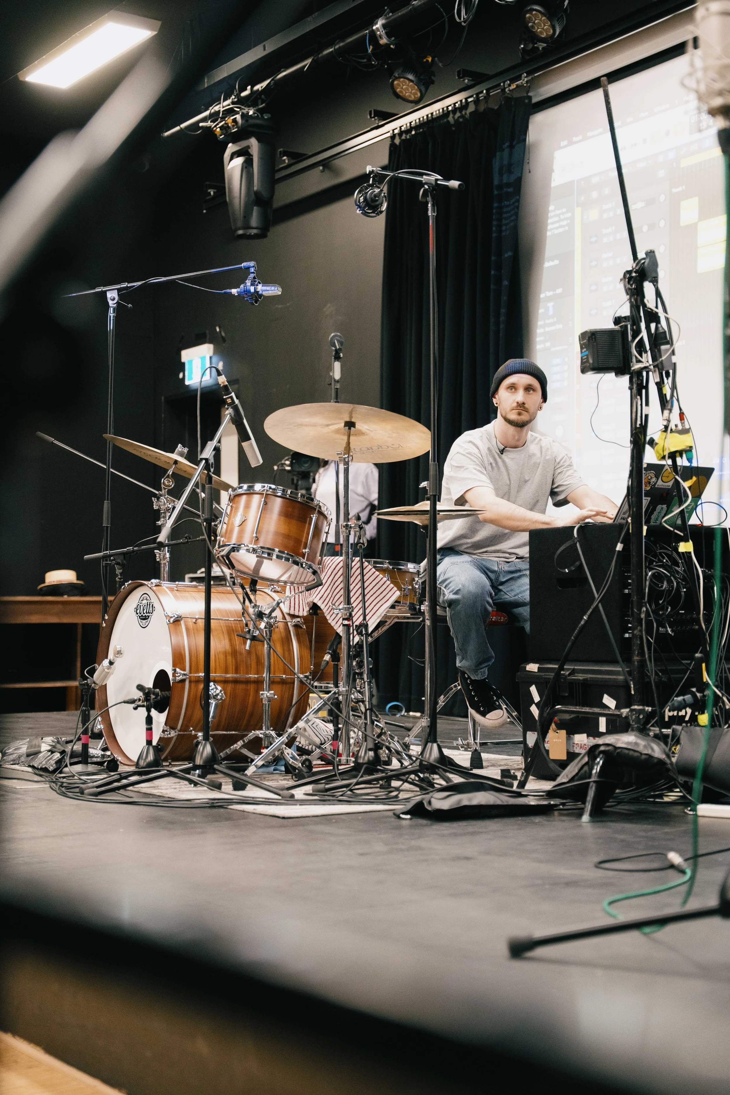 A musician on stage with electronic equipment and a drum set, preparing for a performance in a dark room with black curtains.