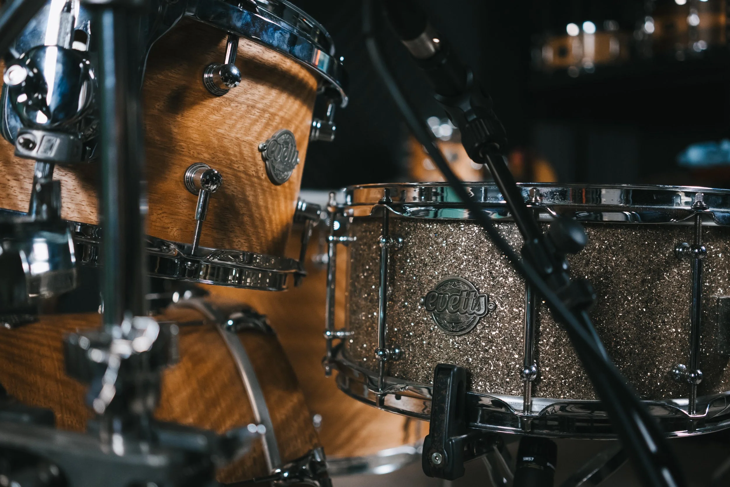 A close-up of a drum set with a wooden bass drum and a glittery silver snare drum, both branded 'Evets', and part of a microphone stand in front.