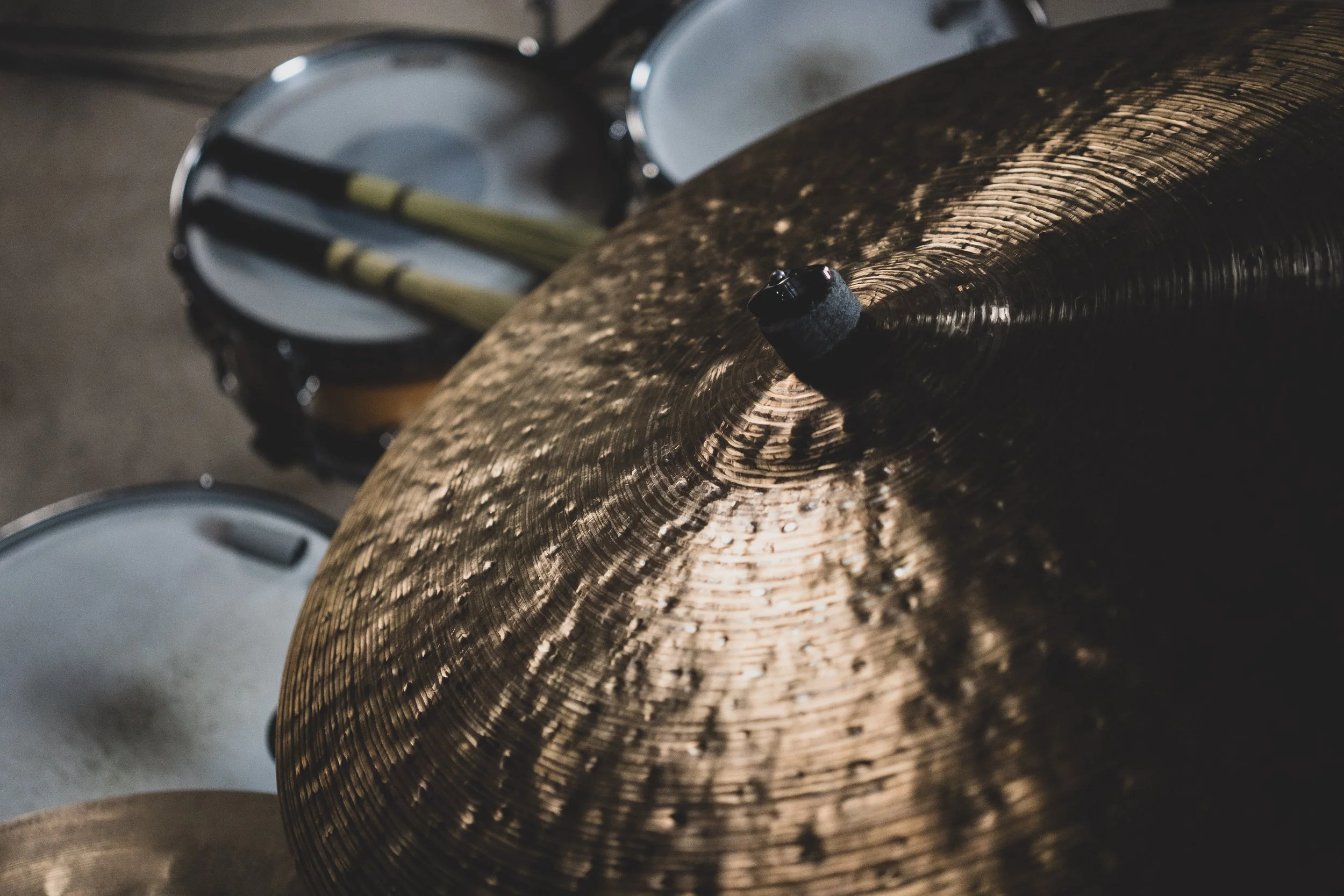 Close-up of a shiny, golden cymbal with a dark mounting hole in the center, with drums and drumsticks visible in the background.