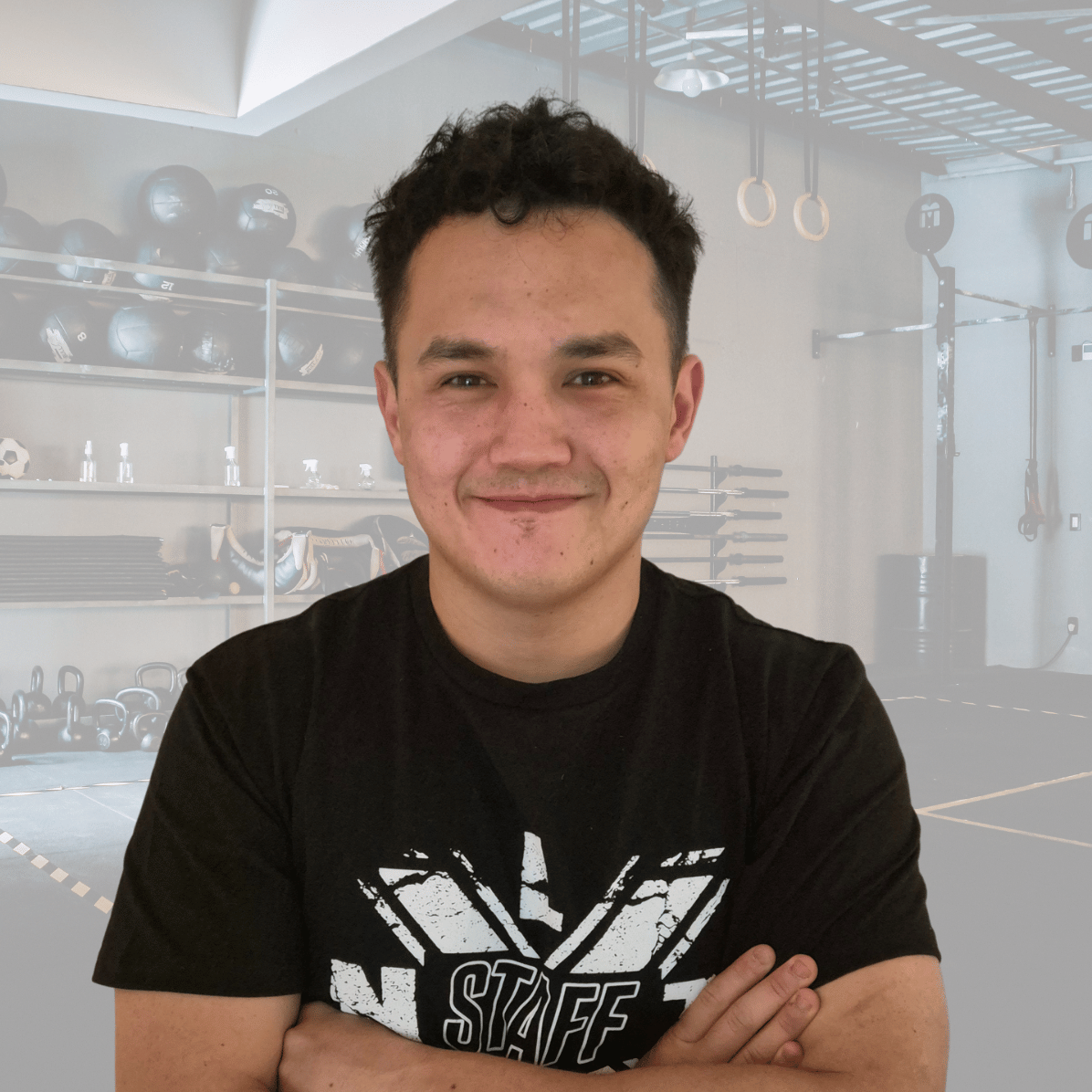 Young man with dark hair smiling and crossing his arms inside a gym with workout equipment in the background.