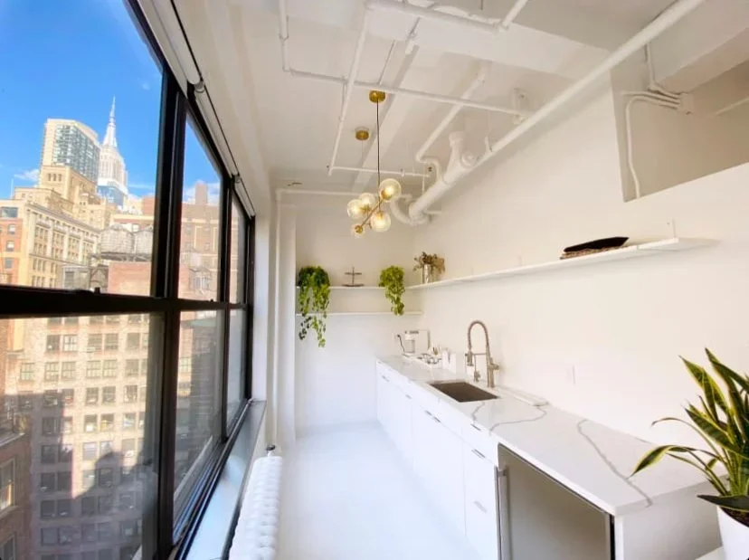 Bright kitchen with large window, white cabinets, marble countertop, black sink, modern gold and glass chandelier, and cityscape view.