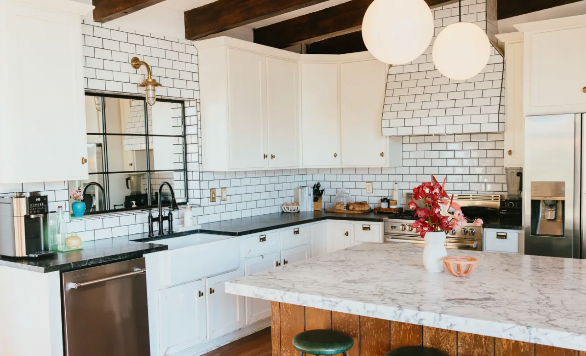 Modern kitchen with white cabinets, black countertops, white subway tile backsplash, and a marble island with a green stool. There are pendant lights, a stainless steel refrigerator, a kitchen sink with black fixtures, and various kitchen items including a flower vase with red flowers on the island.