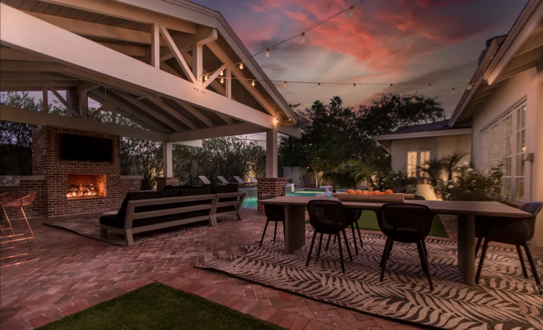 Outdoor patio at sunset with brick fireplace, seating area, dining table with chairs, string lights, pool, and a colorful sky.
