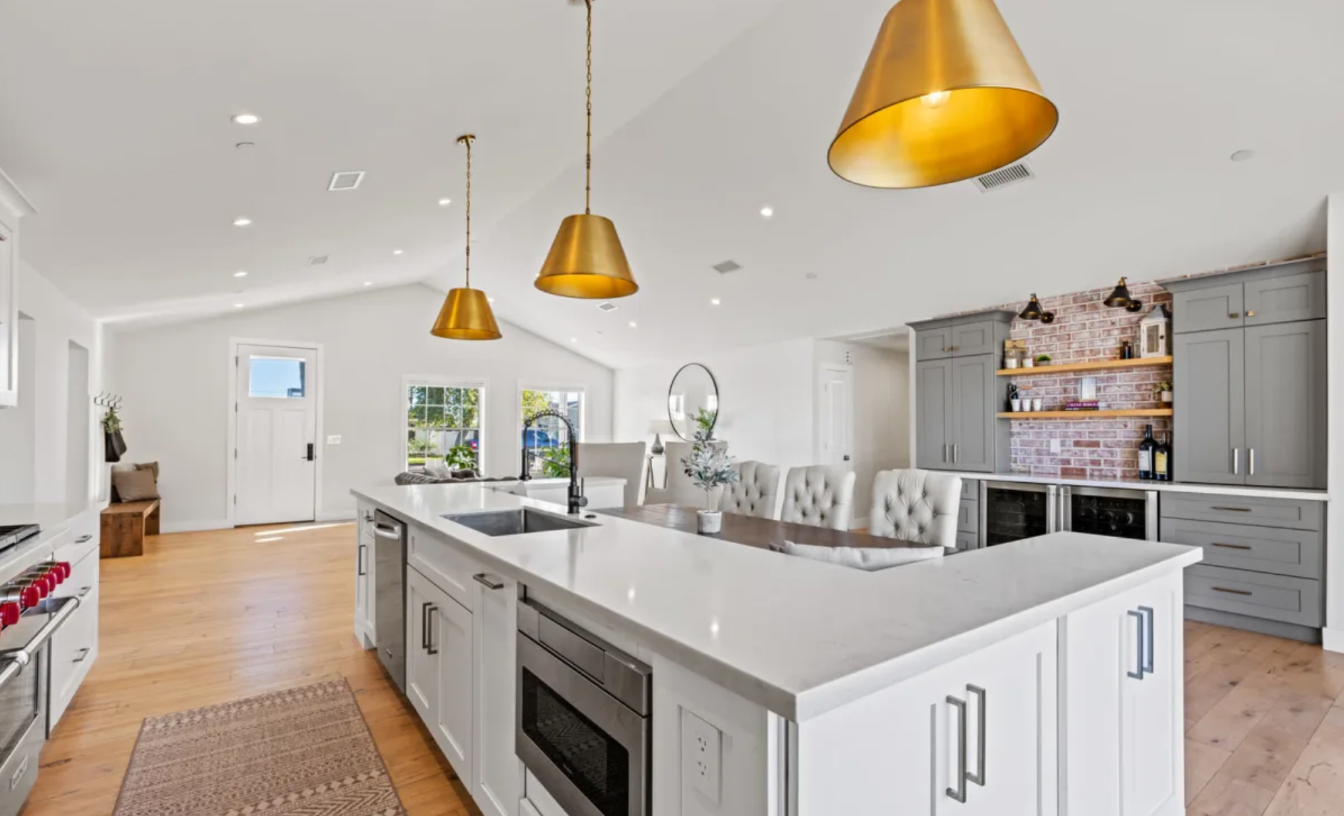 Modern kitchen with white cabinets, a large island with a sink, gold pendant lights, and a brick accent wall with open shelves displaying bottles and decorations.