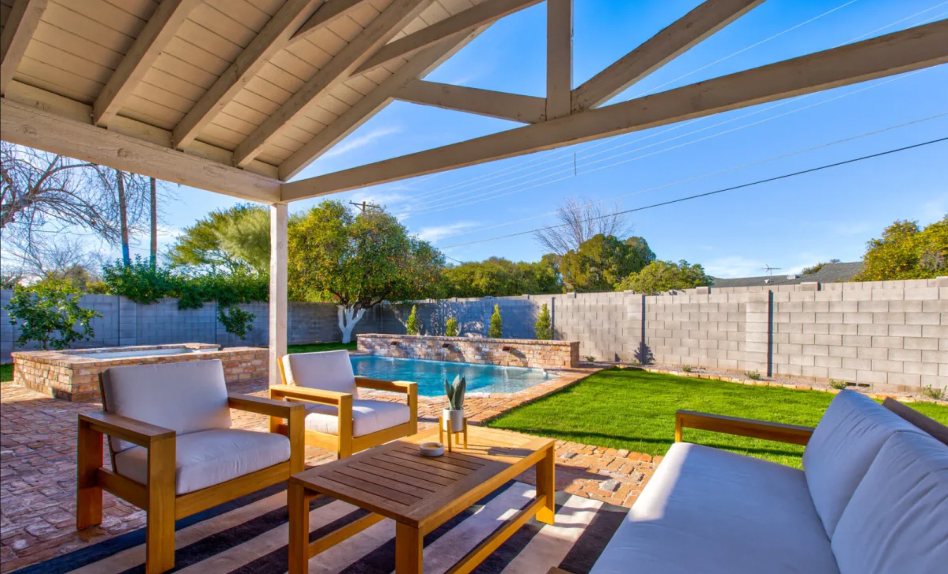 Backyard patio with sitting area, swimming pool, green lawn, trees, and a brick wall under a clear blue sky.