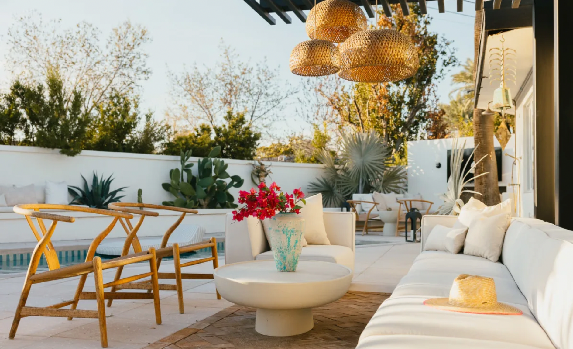 Outdoor patio with white seating, wooden chairs, potted plants, a straw hat on the sofa, and hanging wicker light fixtures