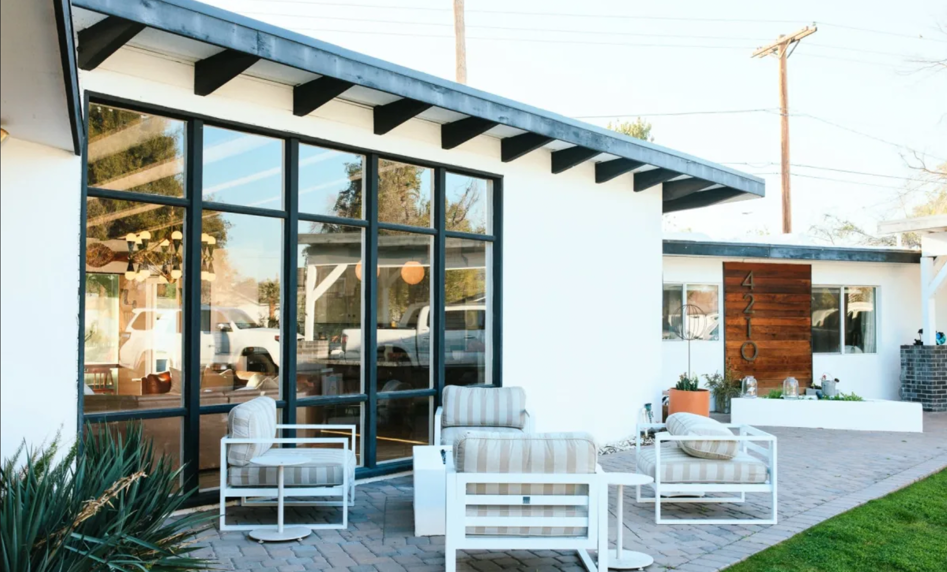 Front patio of a modern house with large glass windows, outdoor seating with striped cushions, potted plants, and a man-sized wooden house number sign
